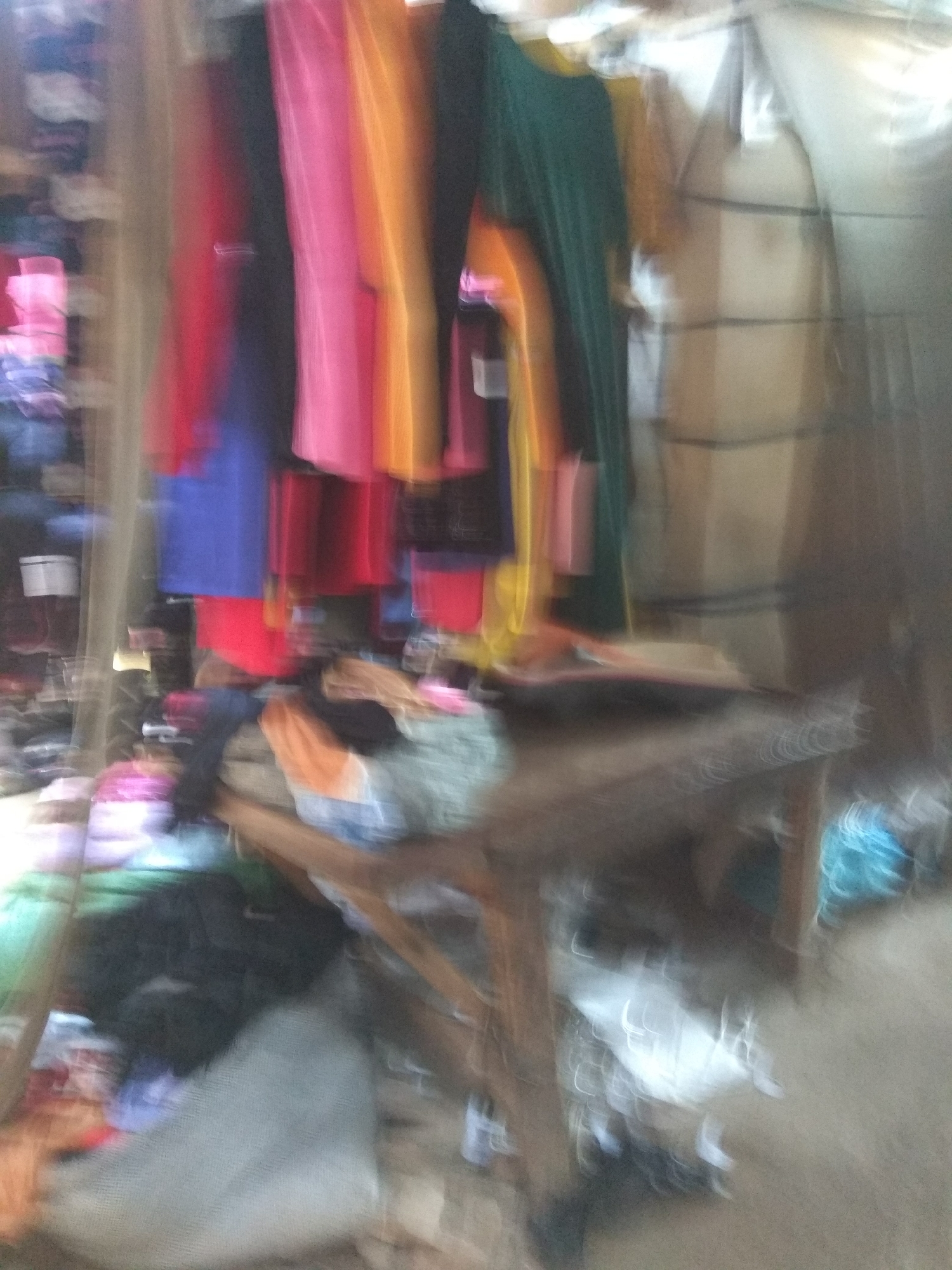 The image depicts a cluttered indoor scene, possibly a market stall or storage area, with an assortment of colorful textiles prominently displayed. The subjects are primarily garments, including what appear to be lengths of fabric, possibly dresses or skirts, in vibrant hues of pink, red, orange, and blue. These are hanging or draped, creating a visually dynamic and somewhat chaotic arrangement. There are also darker, less distinct items that could be clothing or other merchandise piled on shelves and a wooden table in the foreground.
The setting is dimly lit, suggesting it might be late afternoon or evening, or an interior space with limited natural light. The overall impression is one of a busy, perhaps slightly disorganized, commercial environment. The blurriness of the image adds to a sense of movement or perhaps a quick capture, making it difficult to discern specific details or identify any individuals. No text is visible in the image. The presence of textiles in bright colors is a common characteristic of markets in many parts of Nigeria, including Jalingo. The scene conveys a feeling of abundance and variety of goods, typical of a bustling marketplace.