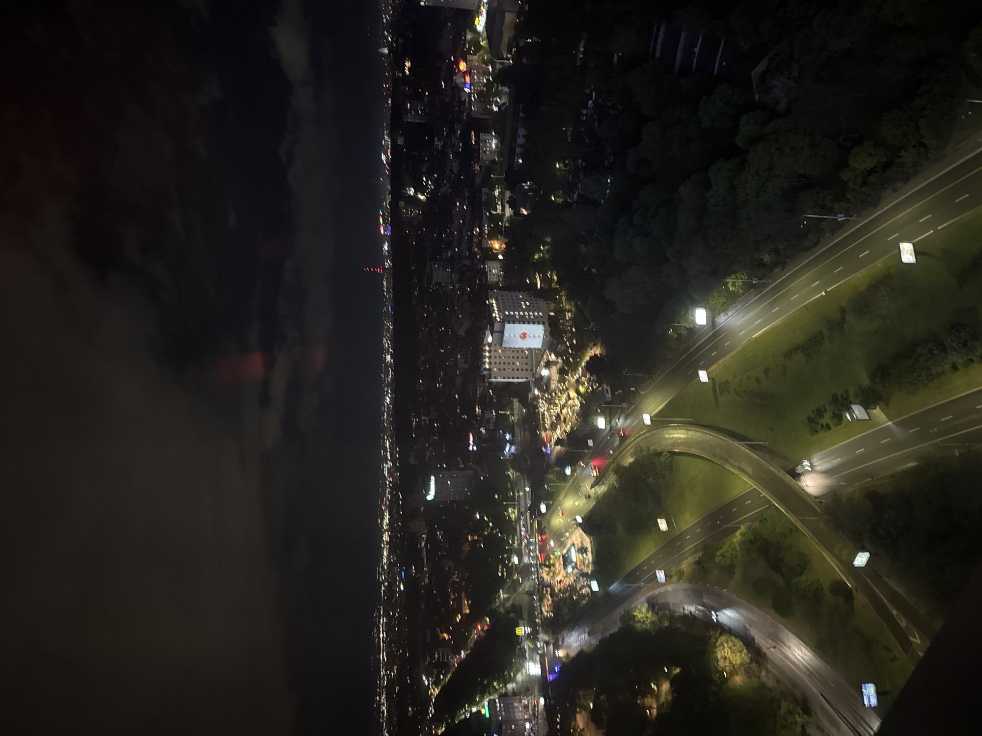 An aerial view, likely from a high-rise building at night, shows the urban landscape of Sofia, Bulgaria. A dark body of water, likely a lake, is visible on the left side, contrasting with the illuminated cityscape. The city's infrastructure, including roads, buildings, and landmarks, are identifiable through their lights. A major highway or road with several lanes cuts through a green area. Buildings with illuminated facades are scattered throughout. A building with a prominent red and white logo or design is identifiable. The overall scene suggests a nighttime urban setting in Sofia.