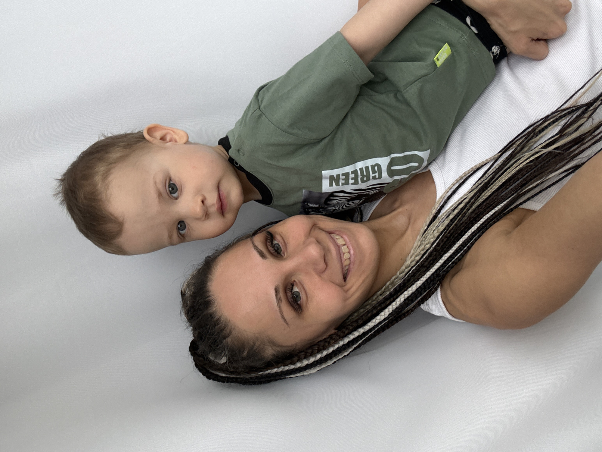 A woman and a young child are lying down together, posing for a photograph in what appears to be an indoor setting with a plain white or very light gray background.
The woman is positioned in the lower right of the frame, smiling broadly with her teeth showing, and looking directly at the camera. She has dark hair styled with numerous thin braids in black, blonde, and white tones. She is wearing a simple white sleeveless top. Her arm is visible, suggesting she is either holding the child or simply lying alongside.
The child, a toddler with light brown hair and striking blue eyes, is lying next to the woman, positioned in the upper left portion of the image. The child also looks towards the camera with a calm, curious expression. The child is dressed in an olive-green long-sleeved top. On the shirt, the word "GREEN" is visible in white letters, appearing inverted due to the child's orientation. A small yellow tag is also present on the side of the child's top.
The overall lighting is soft and even, highlighting their faces and expressions. The image captures a warm and affectionate moment between the two subjects. No specific time of day or weather conditions can be inferred from the indoor setting. The location context for this media file is Rusanovo, Russia.