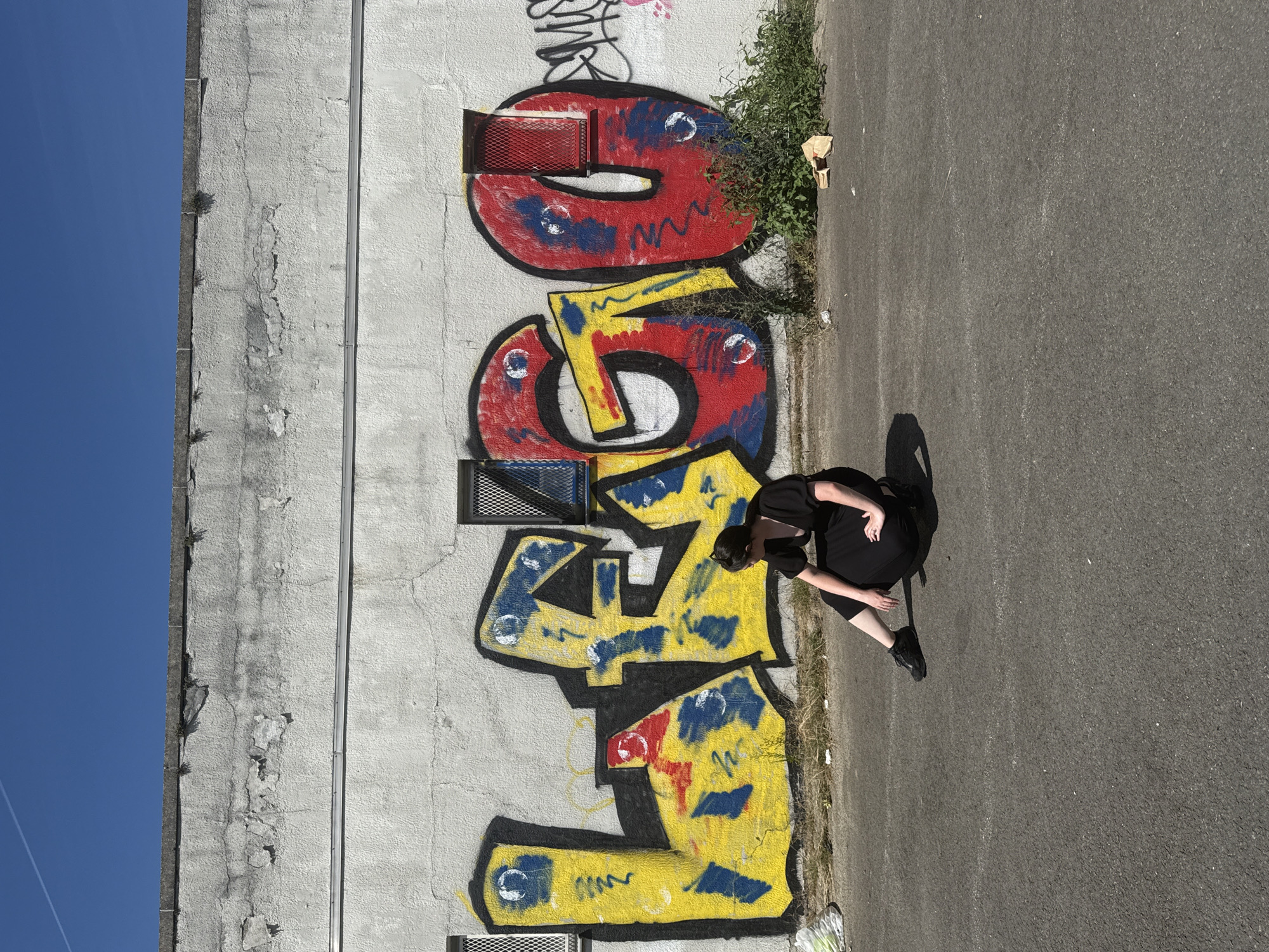 A person wearing a black dress and black shoes is sitting on the asphalt in front of a graffiti-covered wall. The graffiti consists of large, colorful letters that spell out "LEGO". The wall is white and appears weathered, with cracks and peeling paint. There are two windows with metal grates on the wall, one on the left and one on the right. The person is facing away from the camera, looking towards the right. The environment appears to be an urban setting, possibly an industrial area or alleyway, with debris scattered on the ground. The sky is clear and blue.