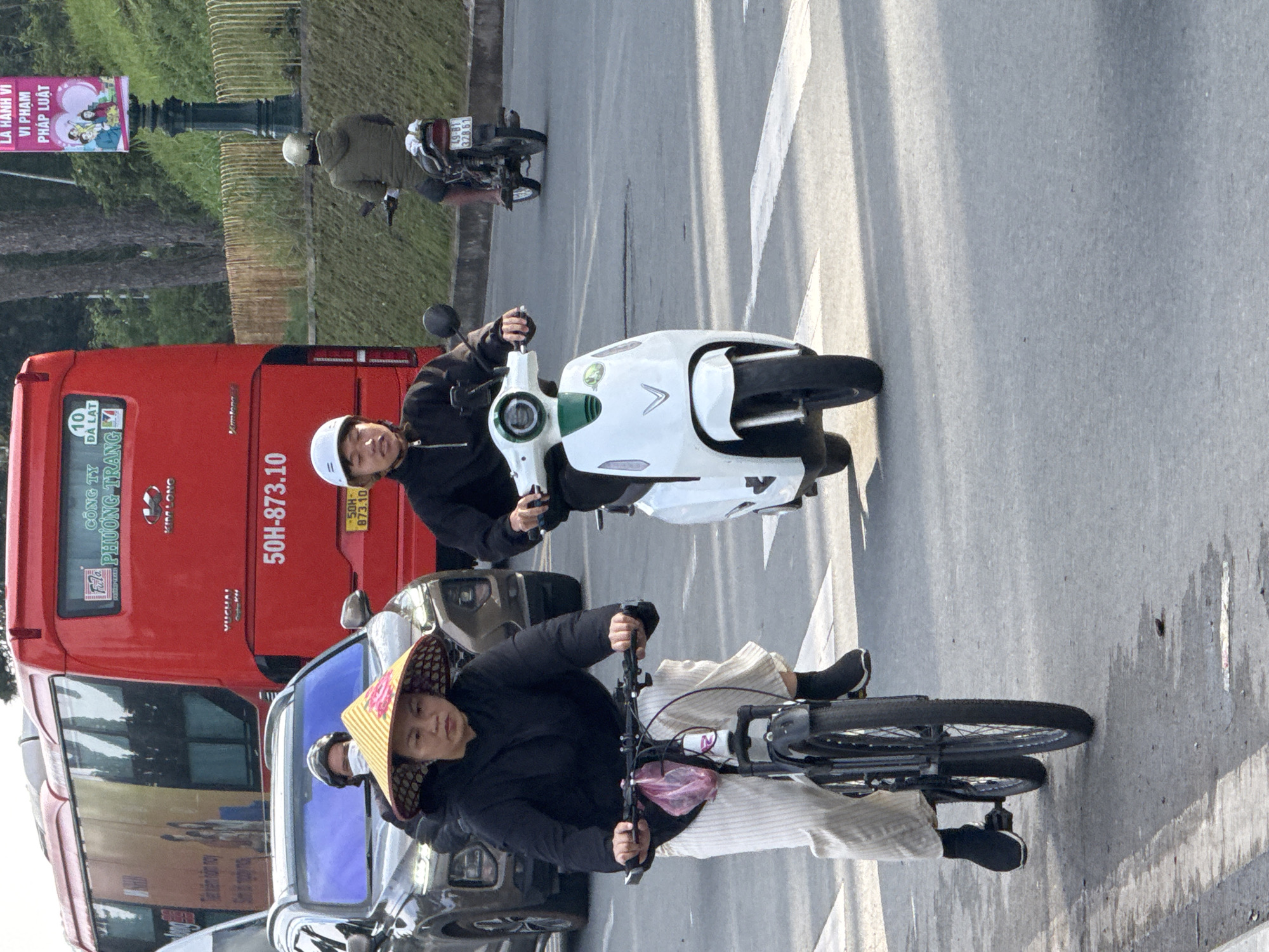 This is a street-level, eye-level photograph taken in Da Lat, Vietnam, likely on a moderately busy street. In the foreground, a woman wearing a conical hat and a black jacket is riding a bicycle. She is dressed in light-colored striped pants and black socks with white shoes. The bicycle is dark in color with white accents. To her right, a white electric scooter is also being ridden by a person wearing a white helmet and a black jacket. In the background, a large red bus with Vietnamese text and signage is visible, along with a dark-colored SUV. Further back, a person on a dark-colored motorcycle is observed. The road surface is asphalt with visible lane markings. The environment appears to be outdoors with trees and a grassy embankment in the background. The lighting suggests it is daytime, with shadows cast on the road. The scene depicts typical urban transportation in Da Lat.