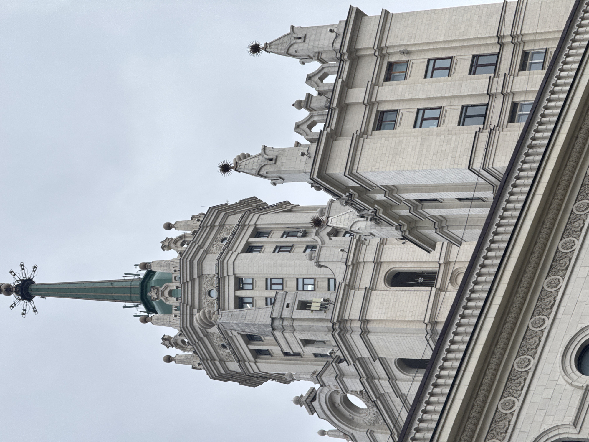 The image shows a low-angle view of a Stalinist skyscraper in Moscow, Russia. The building features a light-colored facade with numerous windows, ornate architectural details, and a prominent spire topped with a star. The sky is overcast.