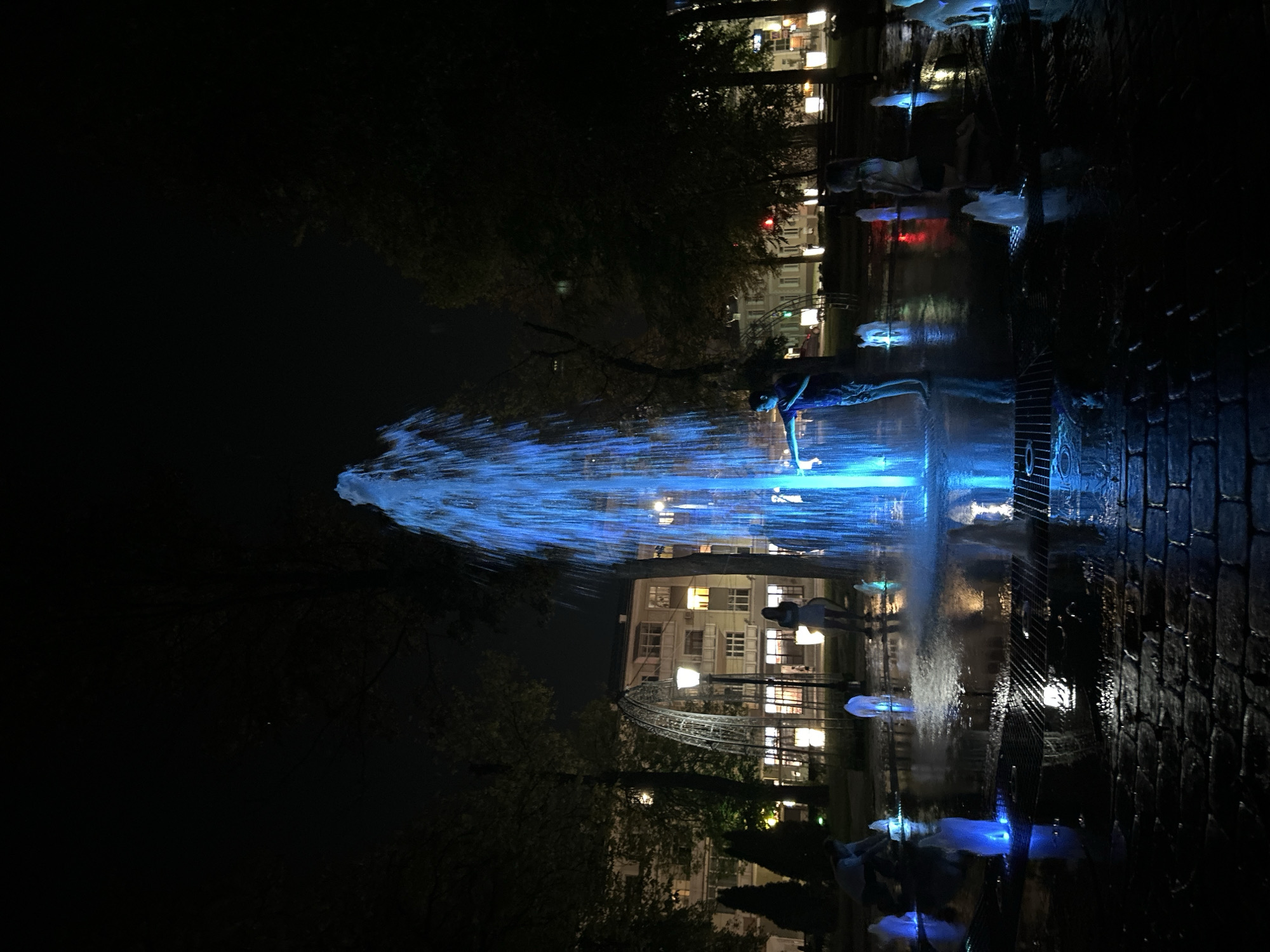 This is a nighttime photograph of a public fountain in the city of Homyel, Belarus. The fountain features a central, tall jet of water illuminated by bright blue lights, with smaller jets of water surrounding it. Several people are visible interacting with the fountain. In the foreground, a person is reaching out towards the main water jet. Further back, several children are playing in the water, some kneeling and others standing. The fountain is located in a paved area with cobblestones, and trees and buildings are visible in the background, dimly lit by streetlights and the fountain's illumination. The overall atmosphere is playful and serene, with the vibrant blue light creating a magical effect against the dark night.