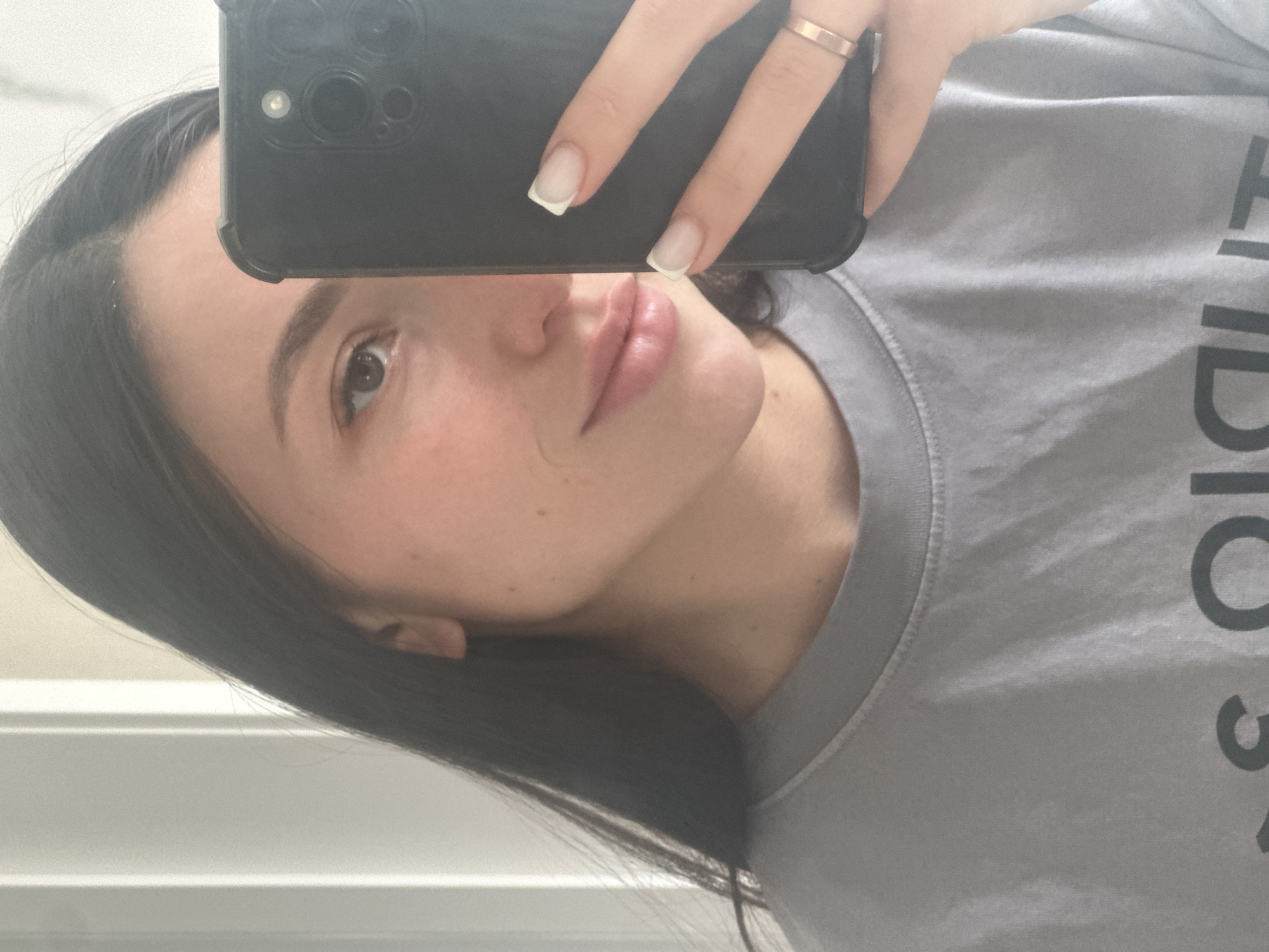 In an indoor setting, a young woman with long, dark hair takes a selfie with her phone. She is wearing a grey t-shirt. The phone, a black iPhone, is held in her right hand, obscuring the right side of her face. Visible on her hand are clean, French-tipped fingernails and a rose gold ring on her right ring finger. Her left eye, eyebrow, and part of her nose and lips are visible. The background appears to be a light-colored wall with vertical lines, possibly indicating a door or partition. The lighting is soft and appears to be natural or diffused artificial light. There is no indication of the specific location within Эстачи, Russia.