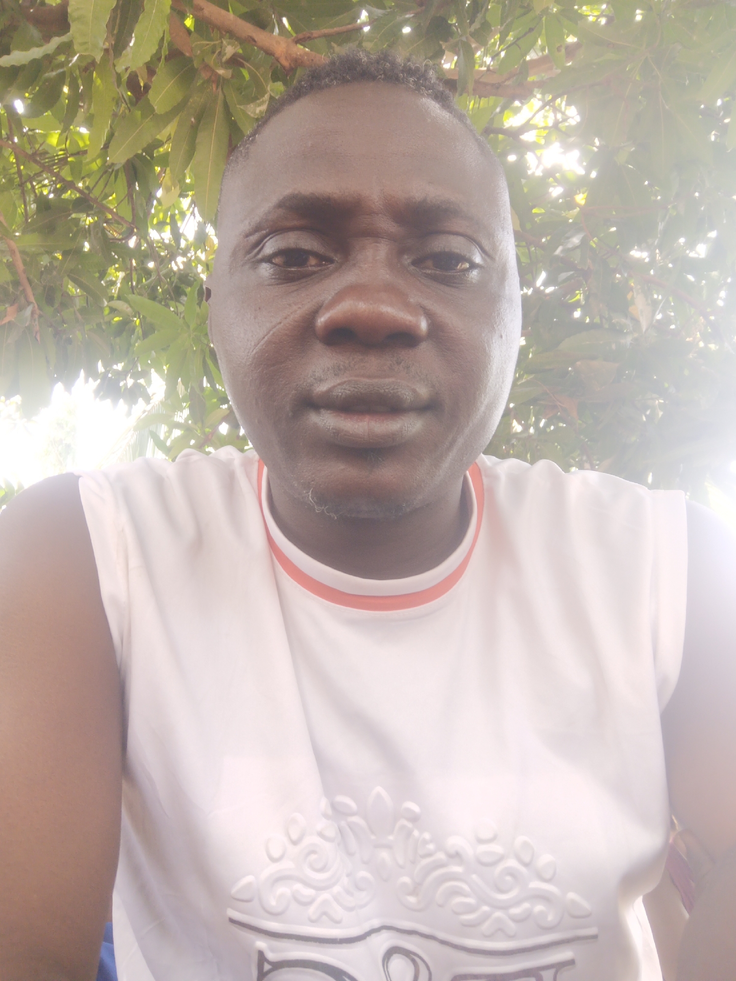 A close-up of a dark-skinned man. He is wearing a white sleeveless shirt with a red-lined collar and embossed designs. The background features green foliage, possibly trees. The image appears to be taken outdoors in natural lighting. The location is Akwanga, Nigeria.