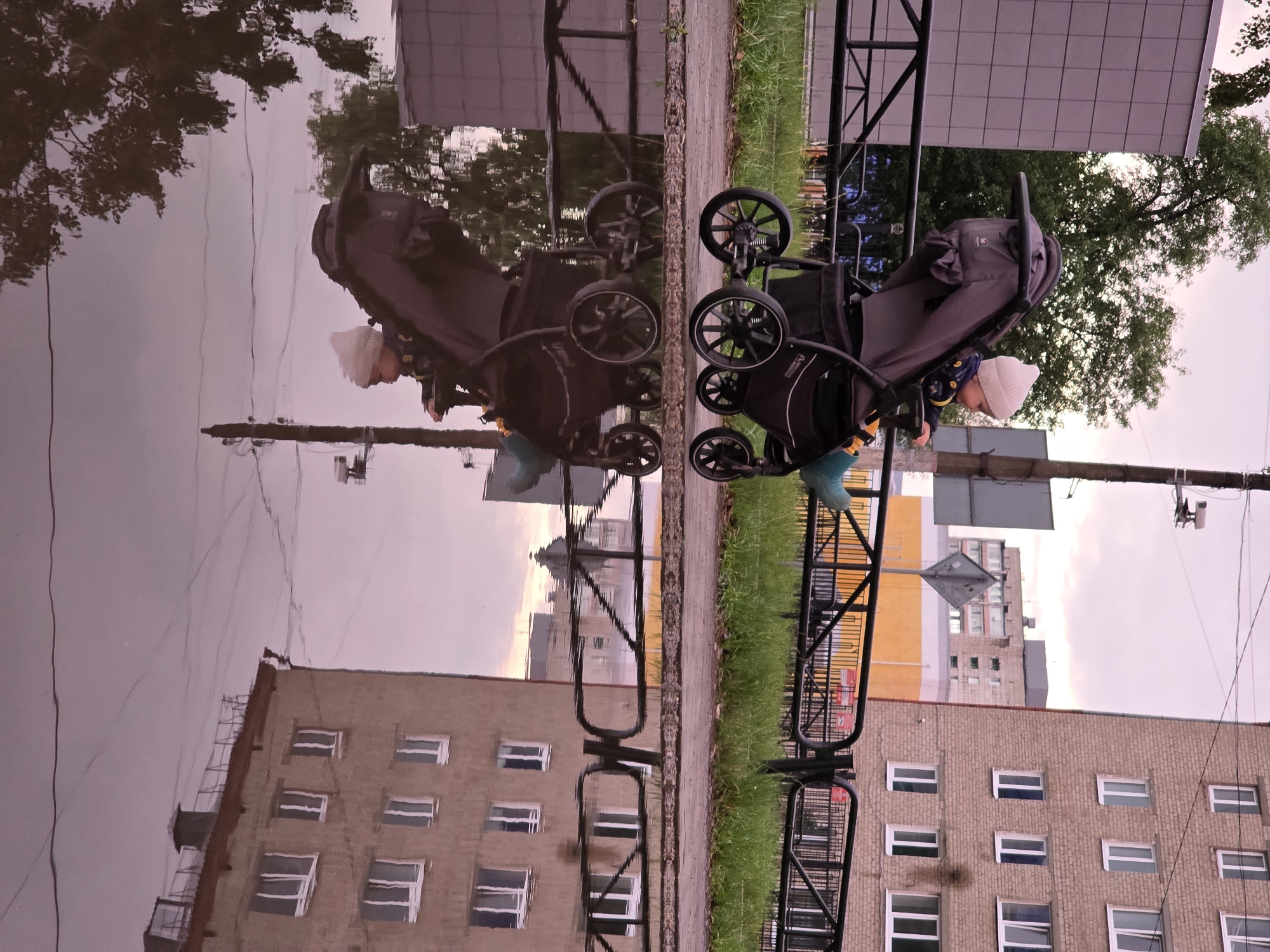 A baby wearing a white hat and a patterned jacket sits in a dark gray stroller on a wet sidewalk. The stroller's wheels are large and black. In the foreground, a large puddle reflects the stroller, the baby, trees, and buildings. The buildings are apartment blocks with multiple windows. One building, constructed of light brown brick, has a reflection showing multiple rows of white-framed windows. Another building in the background is yellow and white. A black metal fence runs along the sidewalk, separating it from a grassy area. The sky is overcast with a soft pinkish hue. The image appears to be taken in the city of Chaikovsky, Russia, based on the architectural style of the buildings and the general environment.
