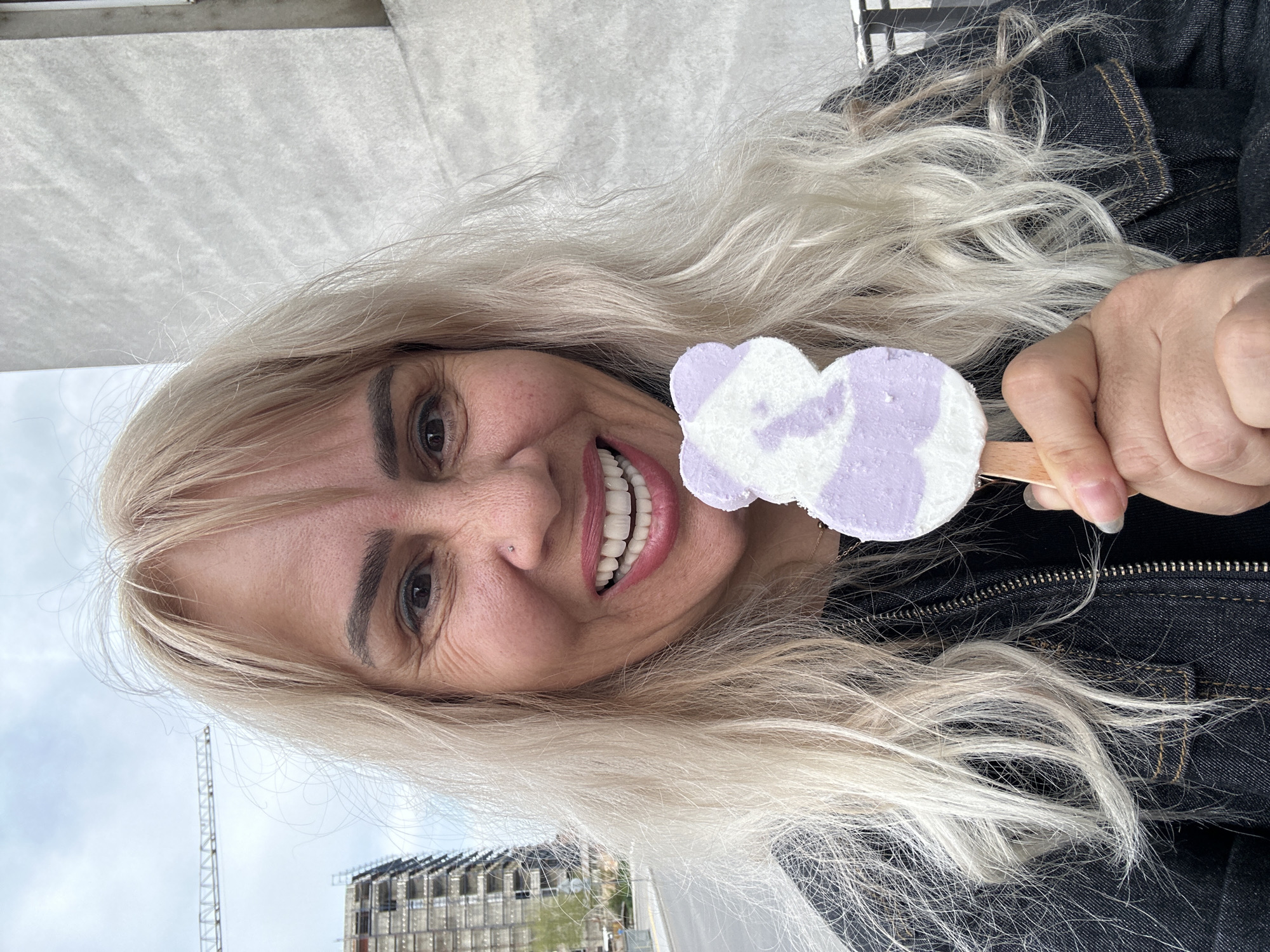 The image, oriented vertically, captures a cheerful woman with long, flowing platinum blonde hair smiling broadly towards the camera. Her dark eyebrows contrast with her light hair, and she has brown eyes. A subtle nose piercing is visible on her left nostril. She is holding a distinctive cloud-shaped popsicle, swirled with purple and white, on a wooden stick, positioning it prominently in front of her.
The woman is wearing a dark denim jacket, visible across her shoulders and arm, with textured fabric and stitching details. Her expression conveys happiness and enjoyment.
The background suggests an outdoor urban setting, likely in Sofia, Bulgaria. Above her (to the right in the original image), a partially cloudy sky is visible. To her left (below in the original image), a tall construction crane and a multi-story building under construction are prominent, featuring scaffolding or an unfinished metal facade. The overall scene suggests a casual daytime moment, possibly under an overcast sky, where she is enjoying a sweet treat. There is no visible text in the image.