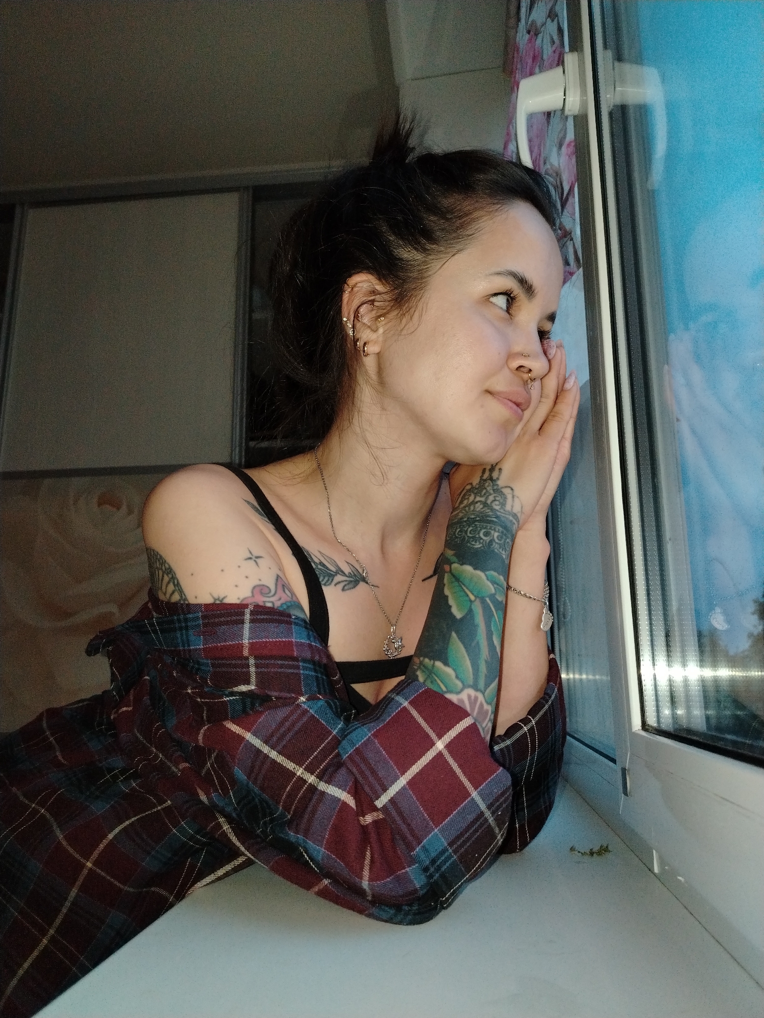 A woman is depicted indoors, leaning against a window, likely in Noviy, Russia. She has dark hair pulled up, fair skin, and multiple ear piercings and a nose ring. She wears a black bra and a red and blue plaid shirt draped off her shoulder, revealing tattoos on both arms and her chest. Her hands are resting on the window pane, and she is looking out of the window with a slight smile. The window frame is white, and the view outside is blurred and blue, possibly indicating night or twilight. The background shows a light-colored wall and what appears to be a cabinet or closet.