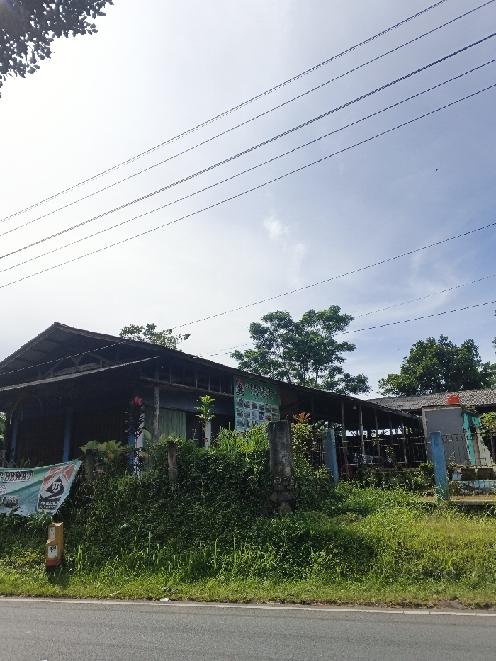 The image presents a daytime scene of a building located alongside an asphalt road, set against a bright, partly cloudy sky. Numerous utility lines stretch across the upper portion of the sky.
The main subject is a rustic, single-story building with a dark roof and light-colored, possibly wooden or painted walls. The structure features open sections, suggesting it might be a small eatery, shop, or a house with an extended porch area. Abundant green vegetation, including tall grass, bushes, and trees, surrounds the building and lines the road, indicative of a tropical environment. A large, leafy tree stands prominently behind the right side of the building.
On the left side of the building, a green banner is displayed, featuring the text "TERA BERAT" and "ANJURAN," along with a logo. Another smaller green sign is attached to the building's wall, partially legible with "KOPI P" visible at the top and several small square images below. A light-colored road marker or post near the left edge of the road displays "KM 101" and "37." A red water tank is visible on the right side of the building. No people are present, and no specific activity is taking place. The scene conveys a tranquil atmosphere under clear daylight conditions, typical of a rural or semi-urban setting.