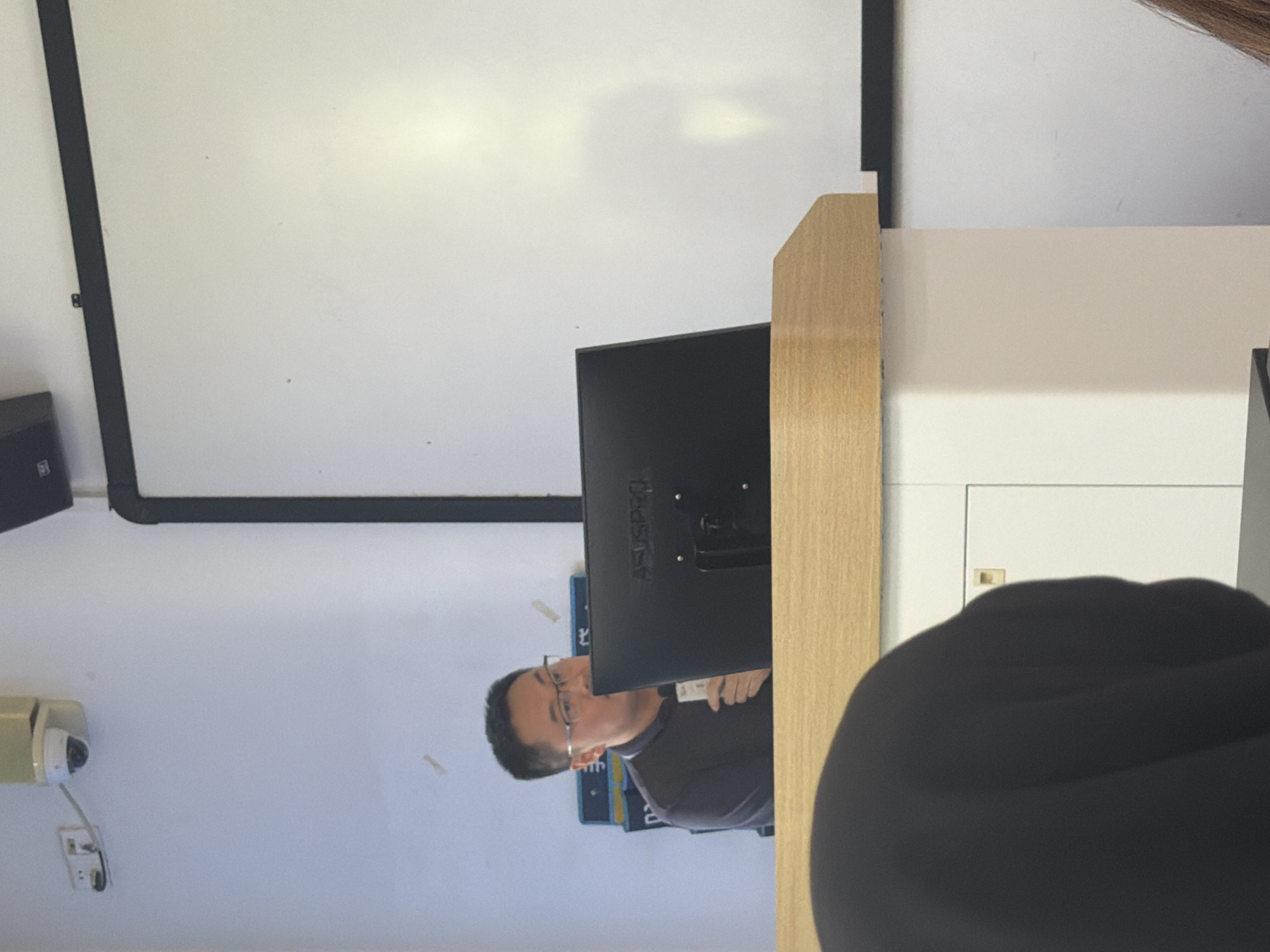 A man wearing glasses and a dark polo shirt is standing behind a wooden podium in a classroom. He is holding a white card in his right hand and looking at a computer monitor placed on the podium. A whiteboard is visible on the wall behind him. A security camera is mounted on the wall in the upper left corner of the image. The setting appears to be an educational institution in Jiangjin, China.