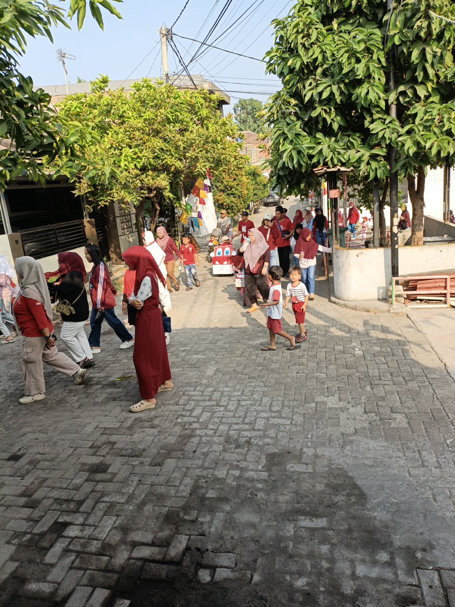 The image captures a street scene in Cilegon, Indonesia, likely during a community event or procession. A group of people, including men, women, and children, are walking along a paved street lined with houses and trees. Many individuals are wearing traditional Islamic attire, such as hijabs and long dresses. Some children are dressed in bright colors, and one child appears to be pulling a decorated cart. The atmosphere seems festive, with colorful flags visible in the background. The sunlight indicates it is daytime, casting shadows on the brick-paved ground. The street is relatively narrow, with houses on either side and overhead utility wires contributing to the urban setting.