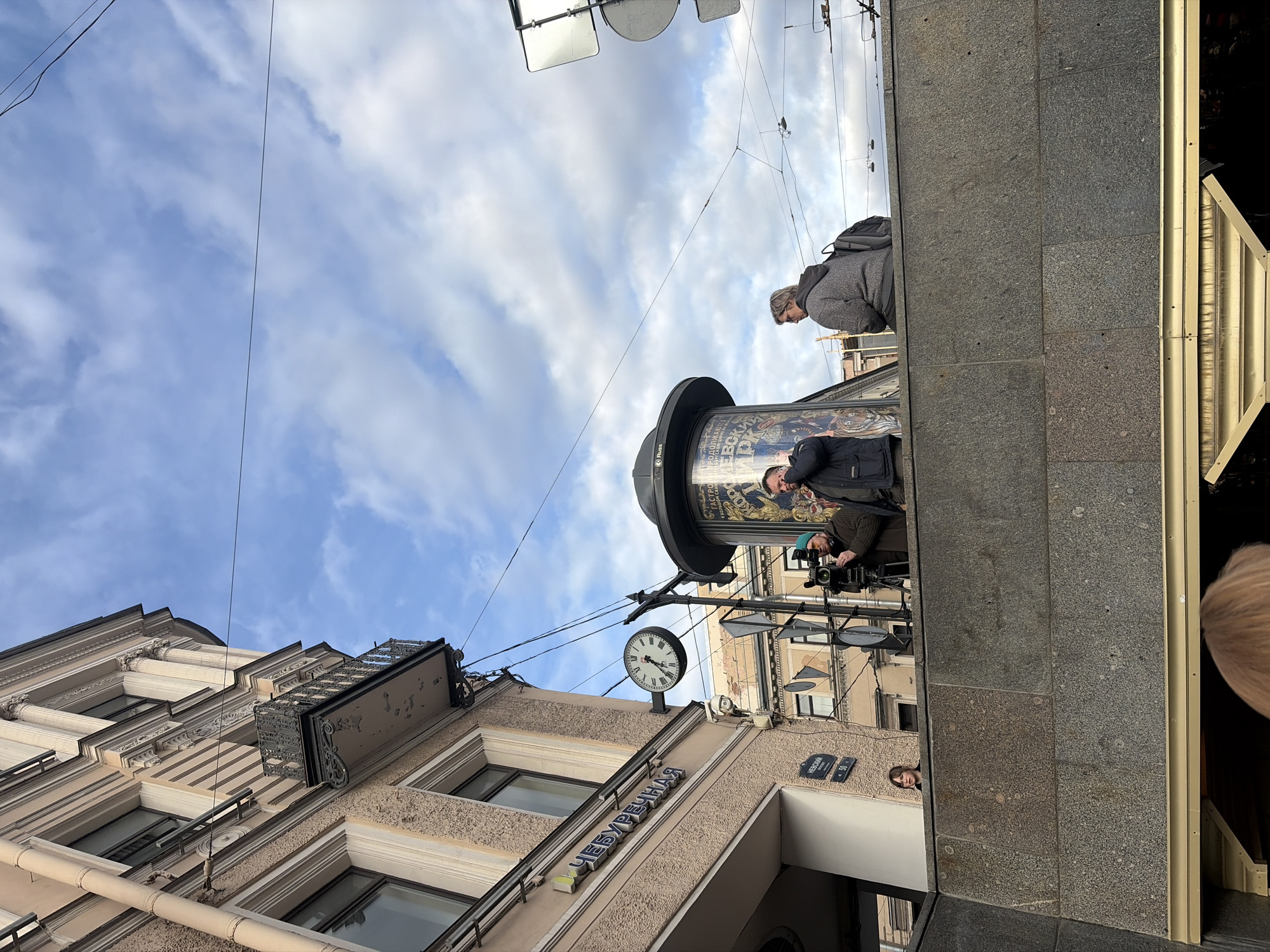 The image depicts a street scene in Saint Petersburg, Russia. Buildings with light-colored facades and ornate architectural details are visible, including windows, a balcony, and decorative trim. Signage in Cyrillic script is displayed on the side of one building. A clock is mounted on the facade, appearing to read approximately 5:15.
A cylindrical advertisement column is present near the buildings. A group of people is situated near the advertisement column. One person is operating a video camera. Another person stands to the side. A woman with a backpack stands nearby.
Overhead, electrical wires crisscross the sky. The sky is partly cloudy, with a mix of blue and white clouds.
A concrete wall is situated to the side, partially obscuring the view. A woman is seen peeking from behind the wall.