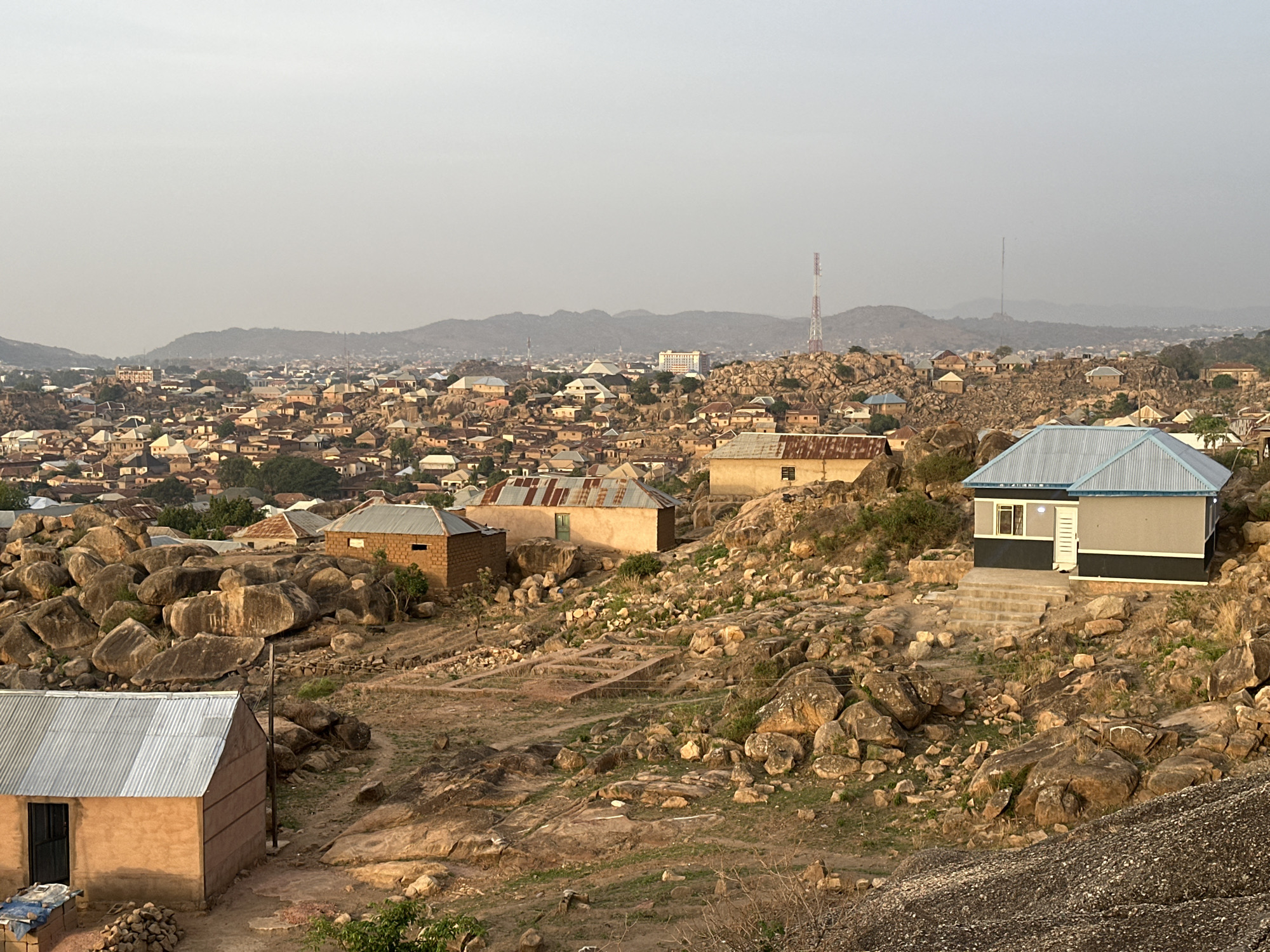 The image depicts a view over the city of Jos, Nigeria, characterized by rocky terrain and scattered buildings. The foreground features several structures with corrugated metal roofs, interspersed among large boulders and sparse vegetation. In the middle ground, a denser area of residential buildings stretches toward the horizon, where hills and communication towers are visible. The sky is overcast, contributing to a muted color palette. The scene conveys a sense of urban development integrated into a natural, rocky landscape.