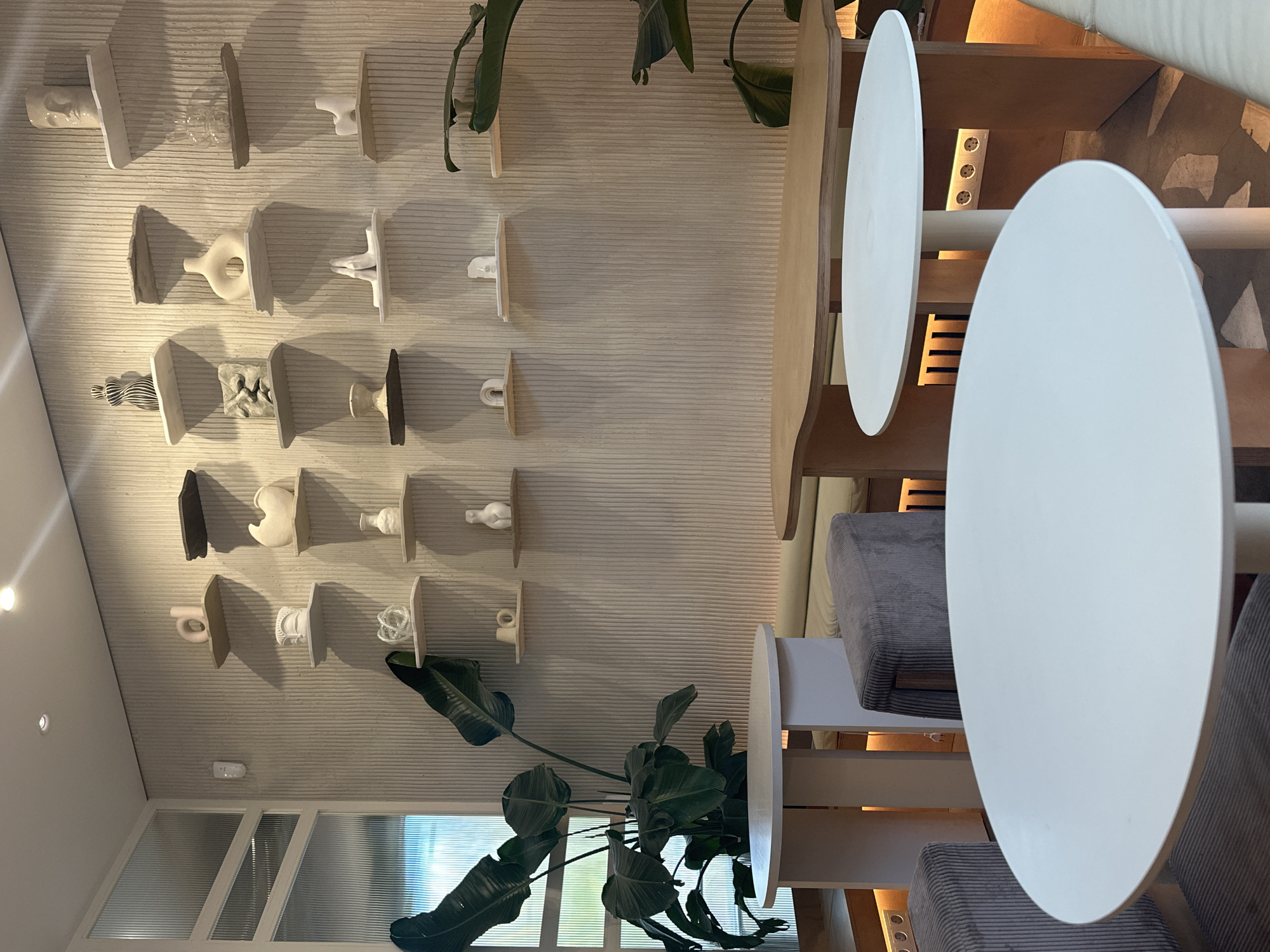 The image captures an interior setting, possibly a café or restaurant, in Novosibirsk, Russia. On the left, the wall features a collection of small, minimalist sculptures displayed on individual shelves. A large, leafy plant stands near the bottom of the frame, positioned in front of a window with translucent glass. In the foreground, two round white tables are prominently featured, each with accompanying chairs upholstered in a grey corduroy fabric. An additional table, made of light-colored wood, is also visible in the scene, along with a multi-socket electrical outlet on the wall. The environment appears modern and design-focused.