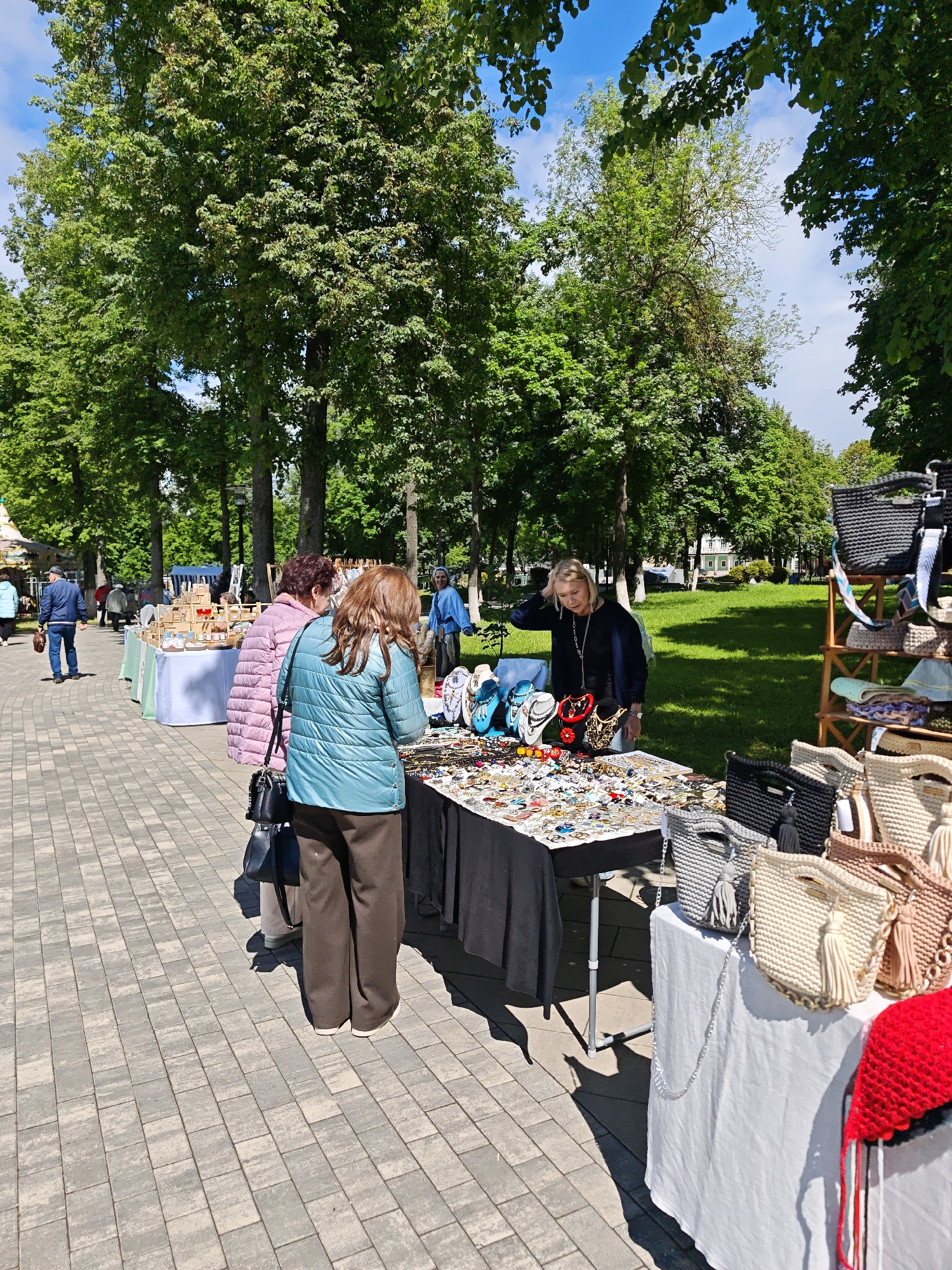 The image shows an outdoor marketplace on a brick-paved area, with trees lining the background, possibly in Chelganovo, Russia. There are multiple tables displaying items for sale. At the main table, draped with a black cloth, a woman with blonde hair stands behind it, interacting with two female customers who are examining jewelry. The woman is wearing a dark-colored top and the customers are wearing jackets, one pink and one teal. Another table is covered with a white cloth, displaying handcrafted bags. In the background, other vendors and customers are visible. A wooden rack displays more handcrafted items. The environment suggests a park or public space, with blue sky visible through the trees.