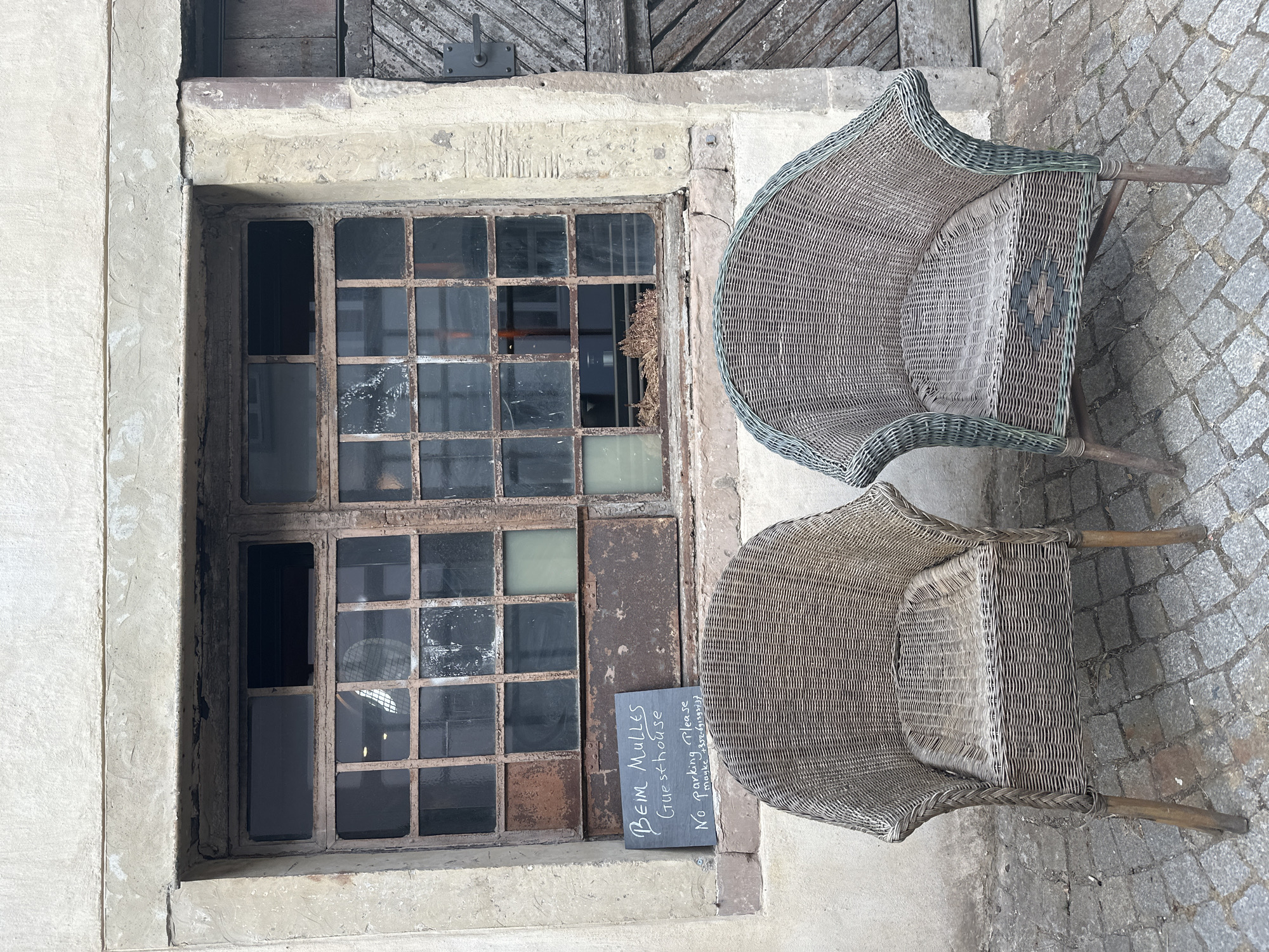 In Vianden, Luxembourg, two wicker chairs are placed in front of a weathered window with a rusted metal frame. A small chalkboard sign rests on the windowsill, displaying handwritten text that reads "BEIM MULLERS Guesthouse No parking Please maybe 0352-691392737". The building's facade is rough concrete, and the ground is paved with cobblestones. A wooden door with a diagonal plank pattern is visible to the right of the window. The scene appears to be an exterior view of a guesthouse.