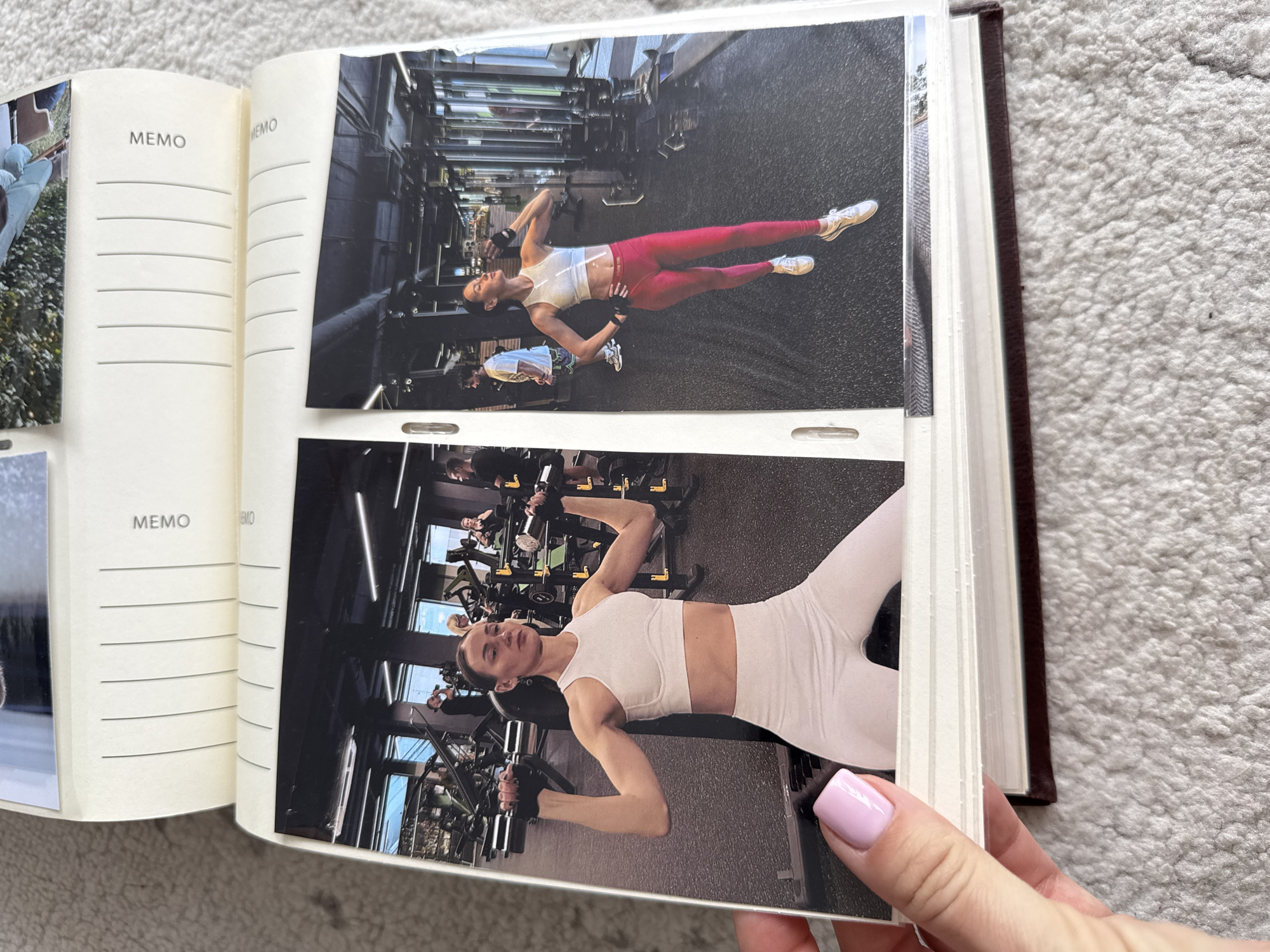 An open photo album is held by a hand with a light pink manicure. The album contains photos of a woman at a gym. In the top photo, she is wearing red leggings and a white top, lying on her back on the floor. A man is standing nearby. In the lower photo, she is wearing white leggings and a matching white top, and is lying on a weight bench lifting weights. The gym is visible in the background of both photos. "MEMO" is printed on the left-hand side of the open album. The album sits on a textured white surface. The location is not identifiable as Sochi, Russia based on the visual information provided.