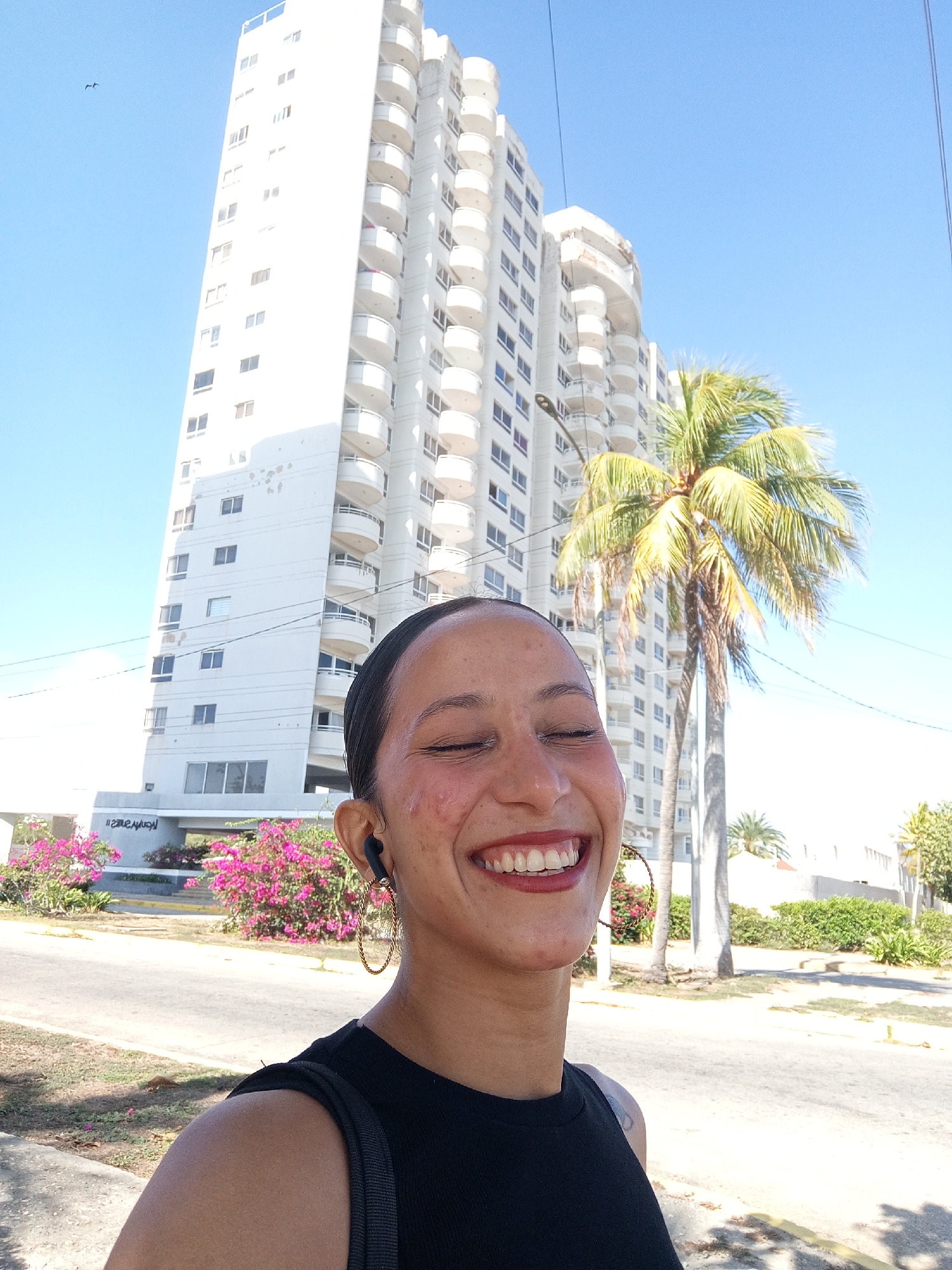 The photograph is taken outdoors on a sunny day in Porlamar, Venezuela, with a building visible in the background. The subject of the image is a smiling female with her eyes closed, wearing a black top and large hoop earrings. She has short dark hair pulled back with a headband and a wireless earbud is visible in her ear. The building in the background is a high-rise with a white facade and protruding balconies. A palm tree is positioned to the right of the building. Pink flowers and greenery are planted in front of the building. The foreground includes a sidewalk and street.