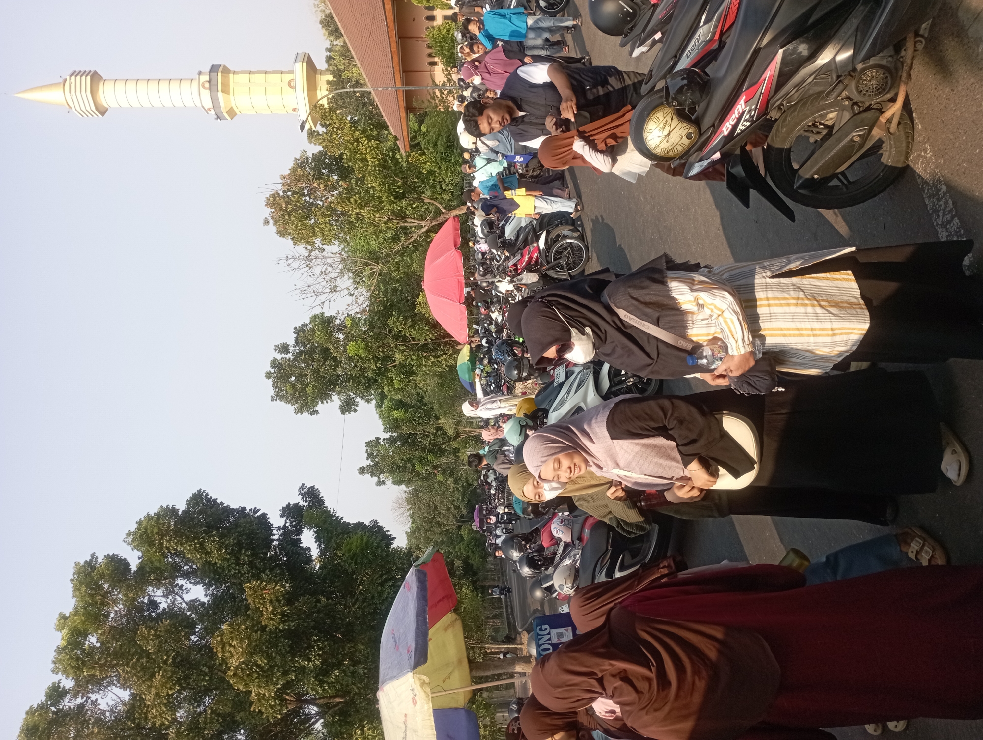 This is a photograph of a crowded outdoor scene, likely a parking lot adjacent to a mosque, given the presence of a prominent minaret in the background. The minaret is tall and yellow, with a pointed golden spire. The surrounding environment includes numerous trees with green foliage, suggesting a warm climate. In the foreground, a large group of people, predominantly women wearing hijabs of various colors and styles, are gathered. Some are talking, others are looking around. Several motorcycles are parked densely in the lot, indicating that most people arrived by this mode of transportation. Umbrellas, one red and one multi-colored, are set up, possibly for shade or as vendor stalls. The ground is paved, and there are signs of a road or walkway. The overall atmosphere appears to be one of a community gathering or event. The image appears to be taken during the day, with sunlight casting shadows. While not explicitly identified, the context strongly suggests a location in Depok, Indonesia, a city known for its many mosques and a population that predominantly uses motorcycles for transport.