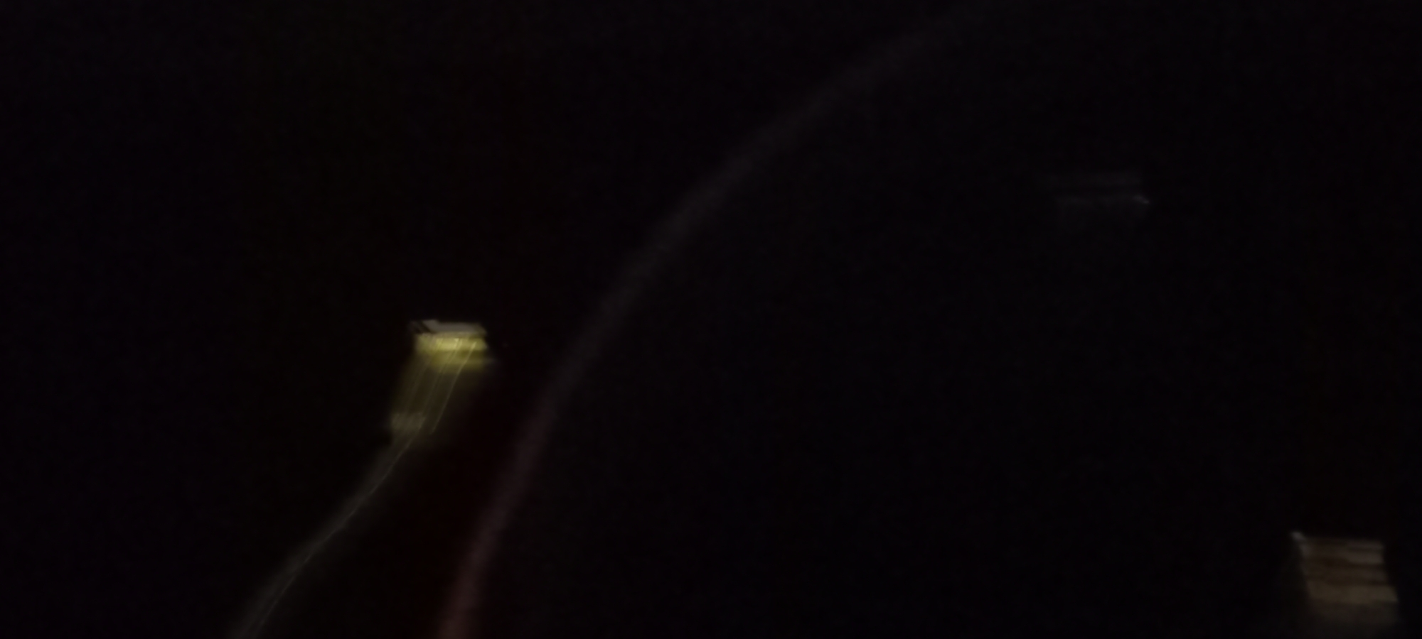 The image depicts a dark, blurry scene with abstract streaks of light. In the upper right quadrant, a rectangular object with a yellowish glow is visible, possibly a light fixture or a distant building illuminated by artificial light. A faint, curved band of light, possibly a light reflection or a spectral phenomenon, stretches across the lower portion of the image. The overall impression is one of motion blur against a very dark background, suggesting a nighttime setting with a moving camera or subject. No specific landmarks or recognizable features of Kryvyi Rih, Ukraine, are discernible due to the low image quality and abstract nature of the content.
