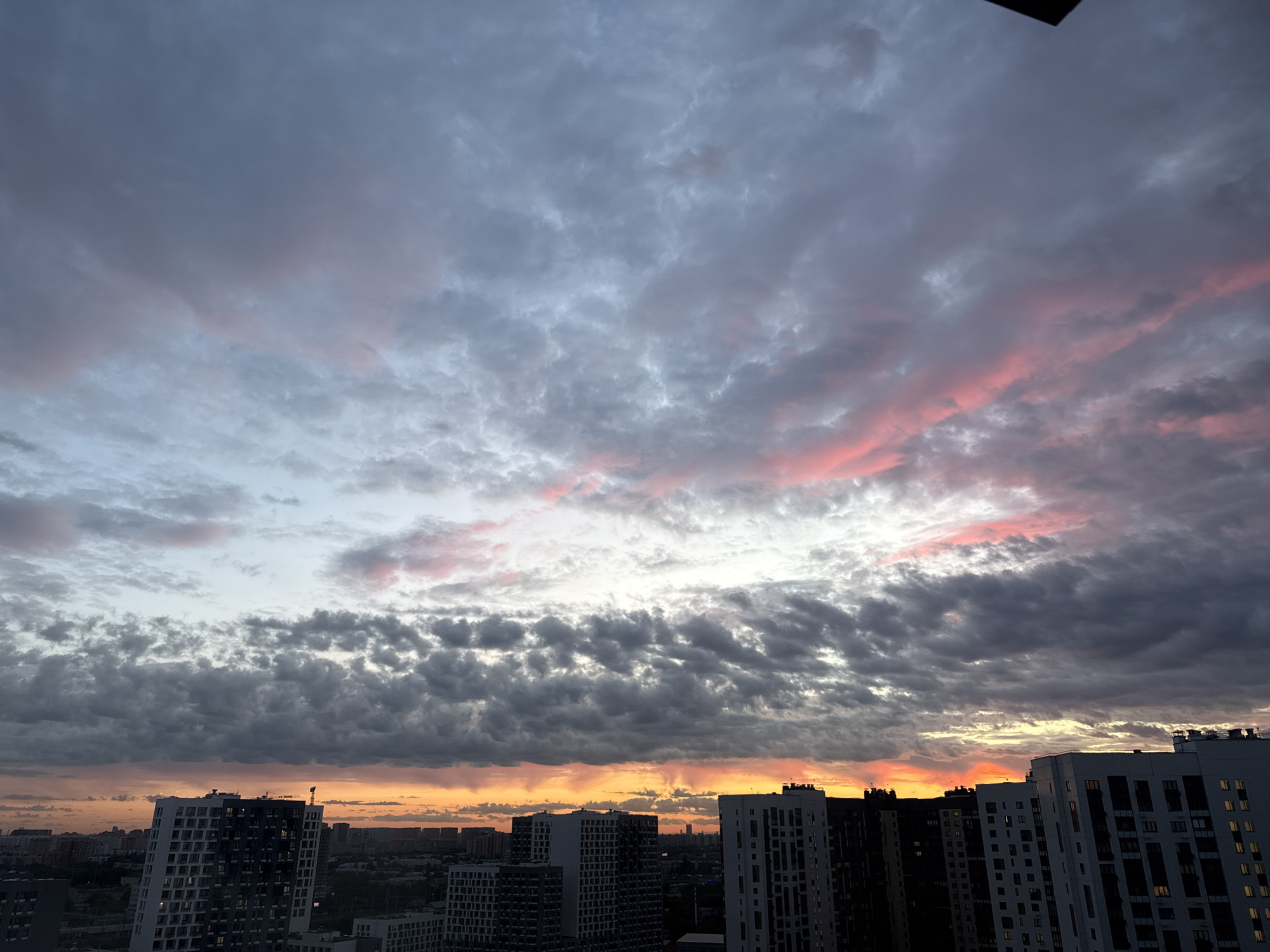 The image captures a city skyline at dusk, likely in Lyubertsy, Russia, judging by the architectural style of the buildings. The sky is dominated by dramatic clouds, with a gradient of colors ranging from deep grays and blues at the top to vibrant oranges and pinks near the horizon, indicating sunset. These colors are reflected on the edges of some of the clouds, creating a striking visual effect.
Below the sky, the silhouette of a modern urban landscape unfolds. Several tall residential buildings, characterized by their multi-story structure and numerous windows, are visible. These buildings are predominantly dark, either due to shadow or their construction materials. Some buildings have a lighter facade, contrasting with their darker neighbors. The buildings are densely packed, suggesting a high-rise urban environment. There are no discernible people or specific objects of interest on the ground. The overall atmosphere is one of a transitional period between day and night, with the natural beauty of the sunset contrasting with the man-made environment of the city.