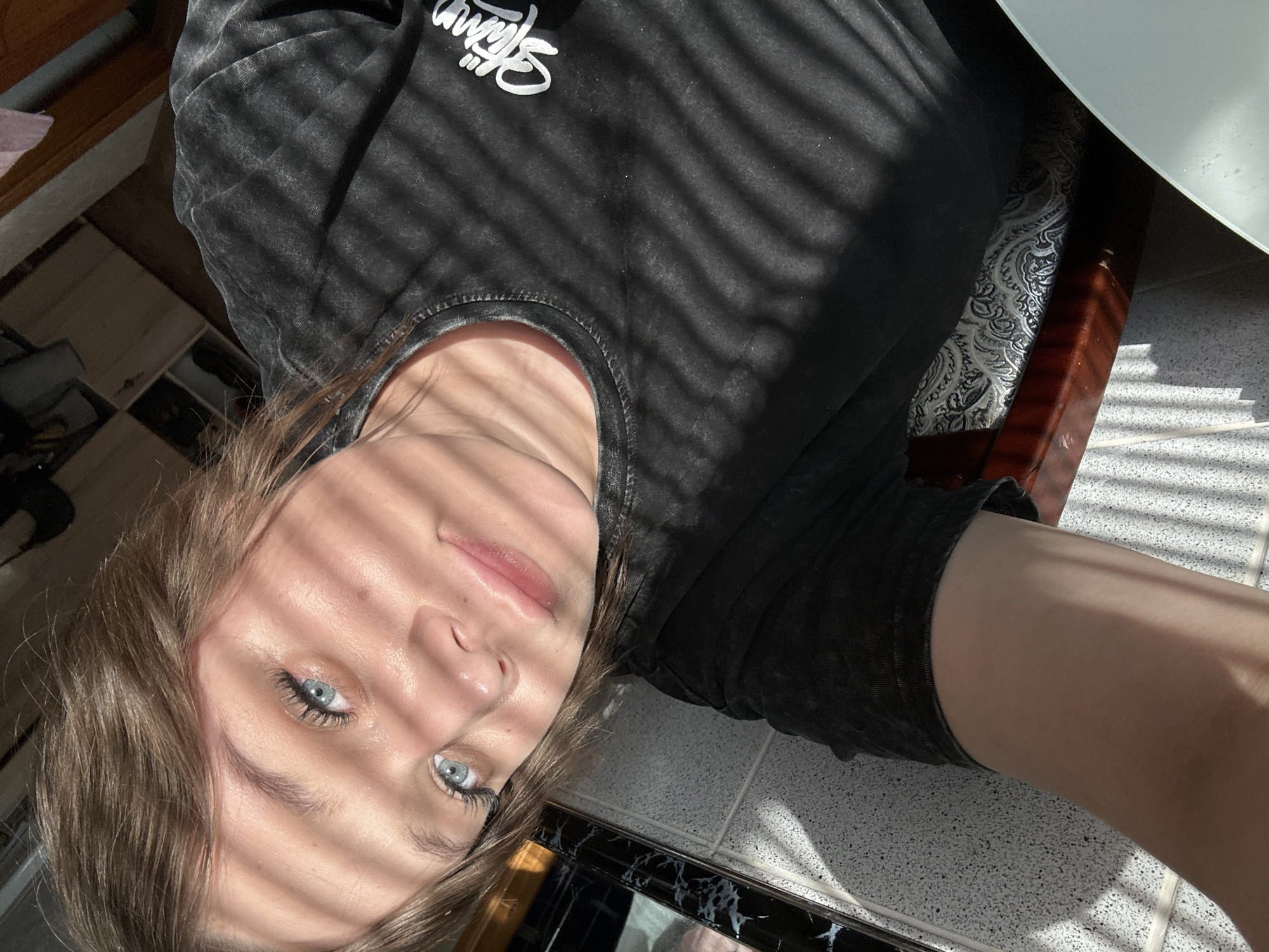 A close-up, angled shot shows a young woman indoors, possibly in an apartment or house, likely in Chaikovsky, Russia. She wears a dark-colored t-shirt with faded writing, which may say "Skyline", and is sitting on a chair with a patterned seat and a dark wood frame, partially obscured by what appears to be a table. The floor is tiled. Light is filtering through blinds, casting distinct shadows across her face and shirt. Her hair is light brown, and she has light blue eyes. The composition gives a casual, selfie-like impression.