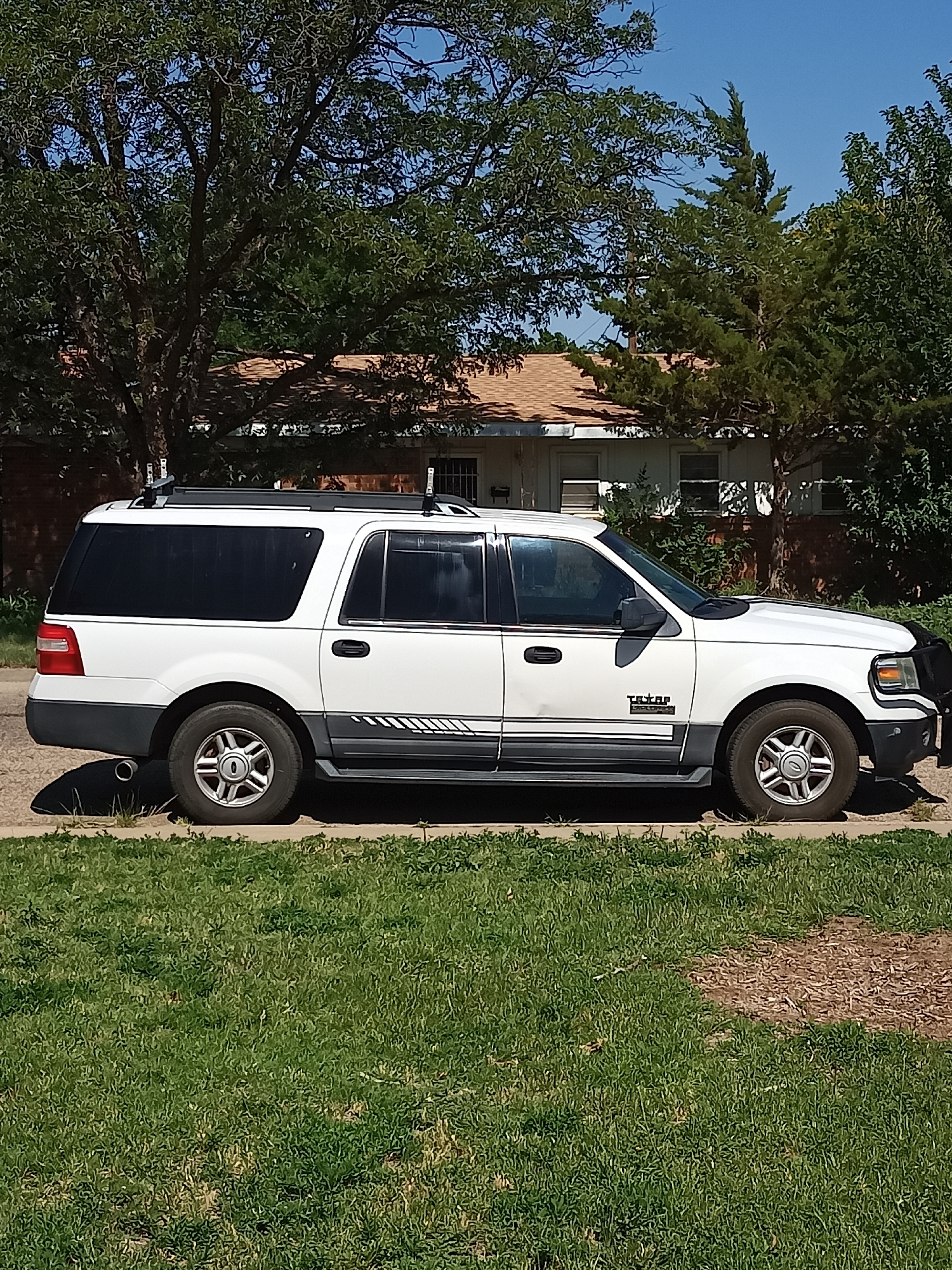 This media file is a full side-view, medium shot of a white, extended SUV parked on a residential street. The SUV is the primary subject, positioned in the center of the frame. It is a sunny day, and the sky is a clear blue. The vehicle appears to be a Ford Expedition, as indicated by the "Expedition" text visible on the side.
The SUV is white with a dark gray lower body trim that includes a decorative stripe graphic. It has tinted windows and a roof rack. The front of the vehicle has a black brush guard. The wheels are silver with a five-spoke design.
The SUV is parked on a concrete surface, likely a driveway or street, with a strip of green grass in the foreground. Behind the vehicle, a single-story house with a brown roof and white siding is visible, partially obscured by mature trees. The trees have full green foliage, suggesting it is summertime or late spring. The overall setting is a quiet, suburban neighborhood.
The image captures a static scene, with no people or obvious activity taking place. The lighting suggests it is mid-morning or early afternoon due to the bright sunlight and distinct shadows cast by the car. The location is identified as Amarillo, United States of America, which aligns with the appearance of the trees and architecture. The scene conveys a sense of normalcy and everyday life in a residential area.