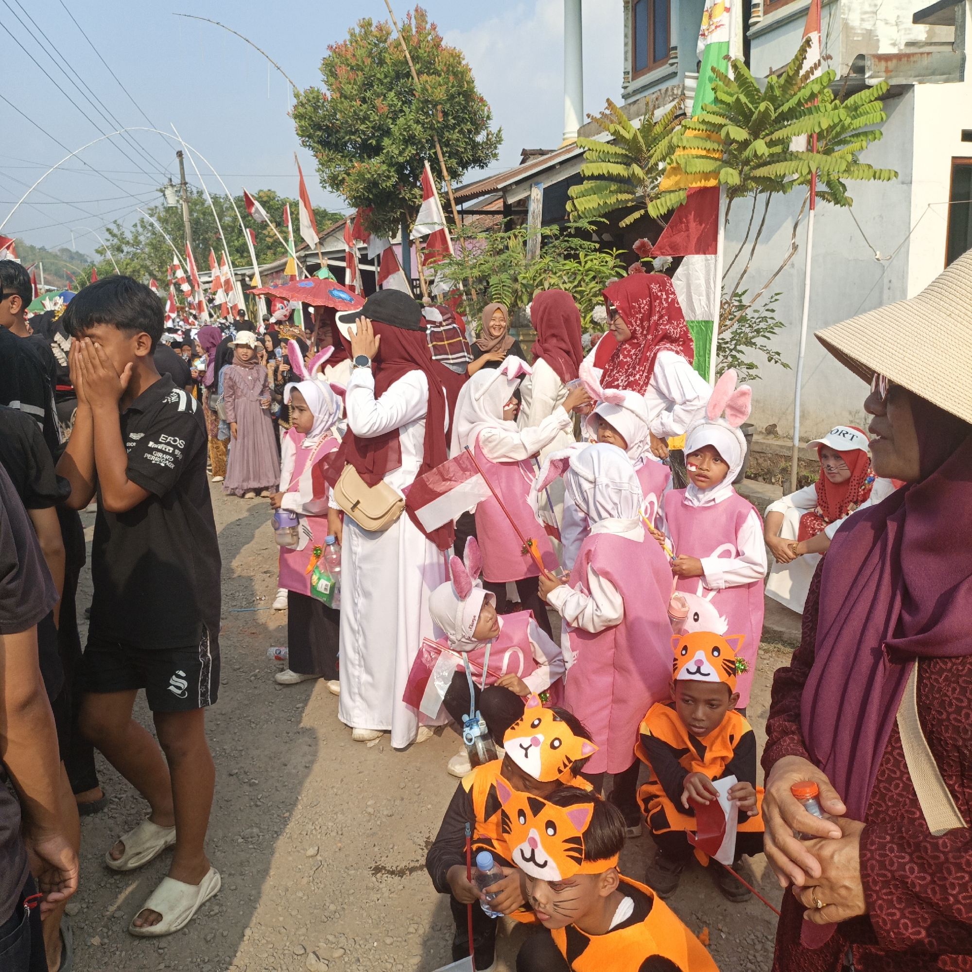 This image depicts a street scene in Tanahbaya, Indonesia, likely during a cultural event or parade, characterized by the presence of Indonesian flags. A crowd of people, including children in costumes, are gathered on a gravel road. In the foreground, a young boy wearing a black polo shirt with white writing is covering his face with his hands. To his right, several children are dressed in pink costumes with white head coverings and bunny ears, some holding small Indonesian flags and toys. Further back, adults are visible, some wearing hijabs and traditional clothing. The environment includes buildings with tiled roofs and vegetation, with power lines crisscrossing the sky. The overall atmosphere suggests a festive and communal gathering.