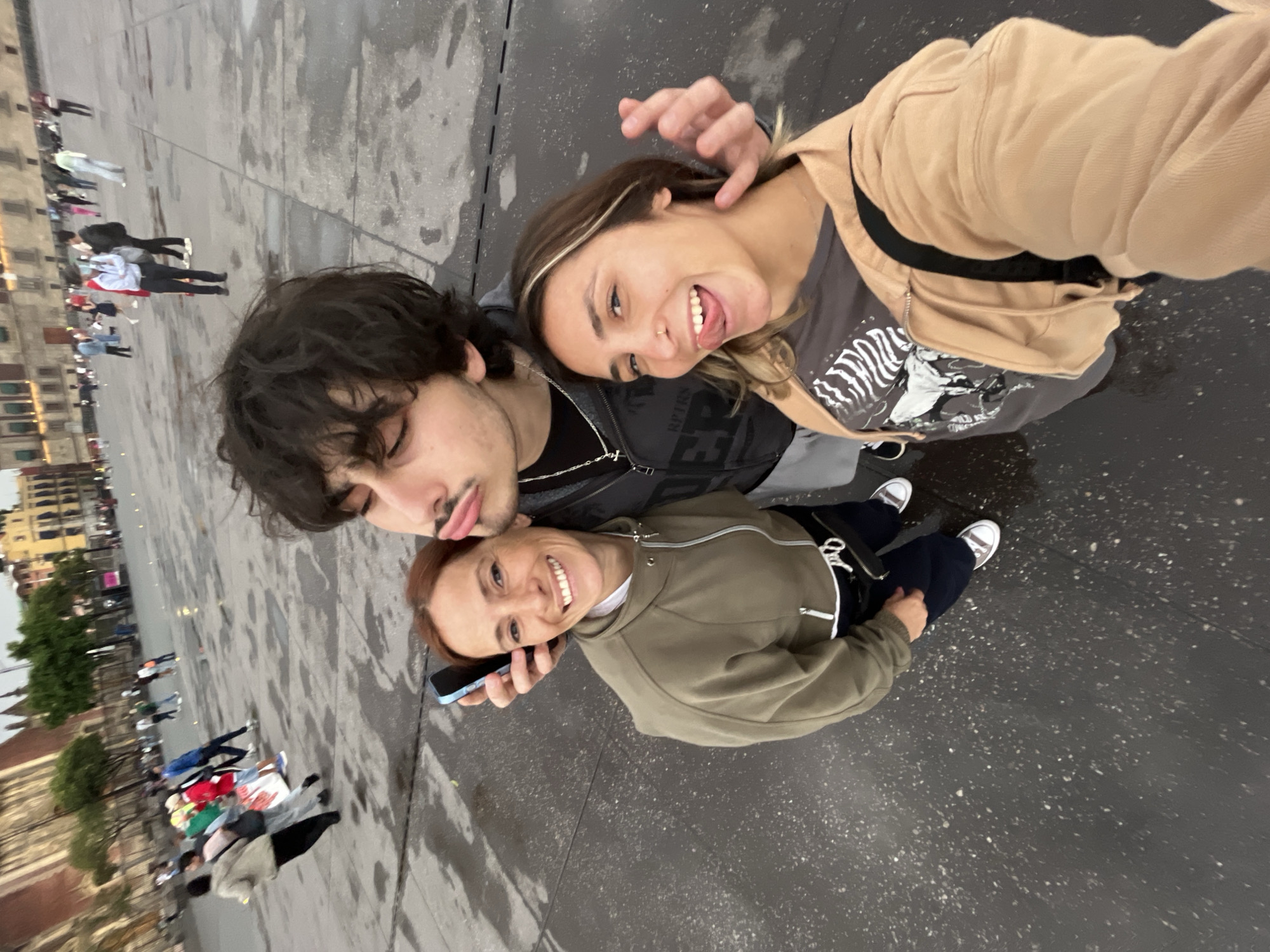 Three people pose for a selfie in Mexico City, Mexico. A young man with dark, shoulder-length hair and a mustache is at the top of the frame, pulling a face. Below him is a young woman with shoulder-length blonde hair and a beige jacket. She has her tongue out. Beneath them is a woman with short, red hair holding a cell phone to her ear. She wears a green jacket. All three are smiling. In the background, people walk across a plaza. To the left are trees and a building. The ground is wet.