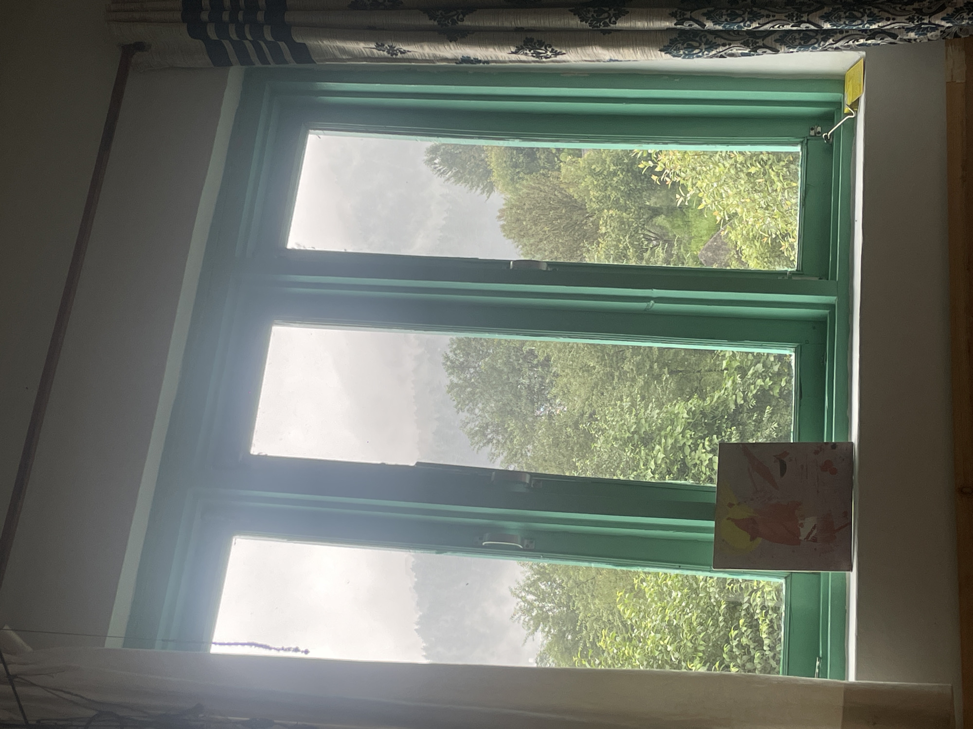 A framed painting sits on a windowsill in Bhagsu, India, overlooking a lush, green landscape. The painting, square in shape, displays abstract splashes of orange and yellow against a muted background. The windowsill is white and appears to be made of marble. To the left of the painting, a cream-colored curtain hangs, partially obscuring the view. To the right, a patterned curtain, with a blue and white damask design, is drawn back.
The window itself is a casement window with vibrant green frames, divided into three vertical panes. Through the glass, a verdant scene unfolds. Dense foliage, a mix of trees and bushes, dominates the foreground. In the background, rolling hills shrouded in a soft, misty haze rise up, suggesting a mountainous region. The sky is overcast, casting a diffused light over the entire scene. The overall impression is one of natural beauty and tranquility.
