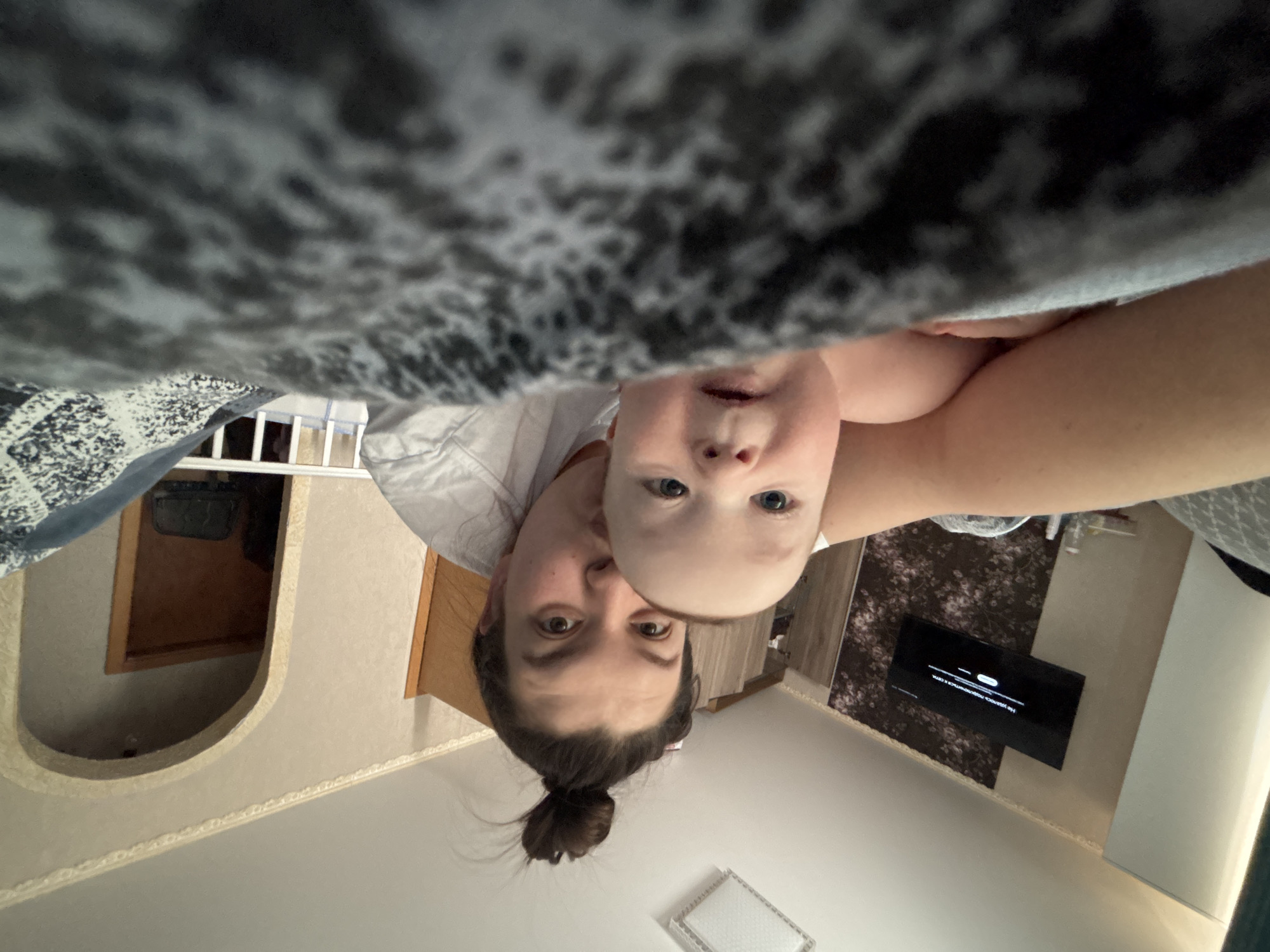 An inverted full-shot photograph captures a woman and a baby looking upwards towards the camera, likely taking a selfie from a low angle. The woman, with dark hair tied in a bun, has a neutral expression, looking directly into the lens. The baby, with light blue eyes and an open mouth, is positioned above and slightly to the left of the woman's face, also gazing at the camera.
The foreground (top of the inverted image) is dominated by a dark, patterned blanket or fabric, partially obscuring the view. In the background (bottom of the inverted image), a home interior in Марковский, Russia, is visible. The room features light beige walls with decorative molding along the ceiling (which appears at the bottom). To the left, a wooden door is set within an arched, textured wall opening. To the right, a flat-screen television is mounted on a wall with dark floral wallpaper. The TV screen displays white text on a black background, which reads "Не удалось подключиться к сети" (Could not connect to the network). A white rectangular light fixture is visible on the ceiling directly below the woman's head. The overall lighting is soft, suggesting an indoor daytime setting.