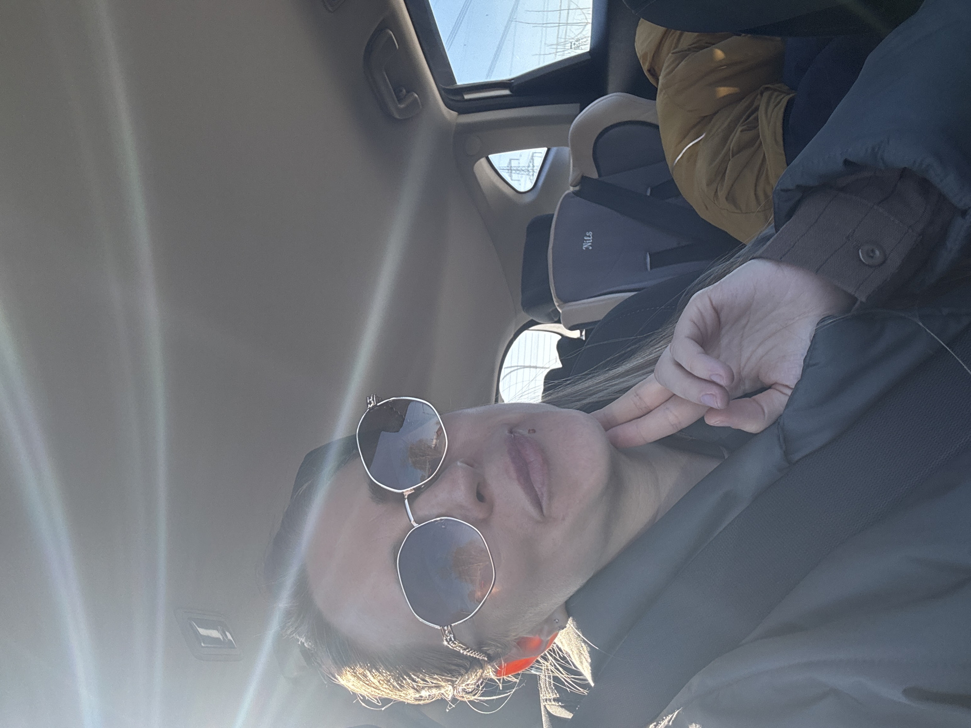This portrait-oriented photograph, taken in Chaikovsky, Russia, captures a person inside a car on a bright, sunny day. The image is taken from a low angle, looking upwards and towards the side of the car. The primary subject is a person, likely female, whose head and upper torso are visible in the foreground, positioned towards the bottom right of the frame. They are wearing fashionable round sunglasses that reflect the bright sky, a dark jacket, and have light-colored hair which glows in the sunlight. A philtrum piercing is visible above their upper lip, and an ear piercing is on their tragus. Their right hand is casually placed near their neck/jawline.
Strong sunlight streams into the car from the left, creating prominent lens flares across the left side and upper portion of the image. The car's light-colored ceiling is visible, extending towards the upper left. An open sunroof reveals a clear blue sky with distant power lines or electrical structures. In the background, behind the person, a grey child safety seat or booster seat is clearly visible, with the name "Nils" printed on its side. A yellow or gold puffy jacket is partially seen next to the child seat. Other car interior details, such as ceiling handles, are also apparent. The scene depicts a moment during a daytime journey or stop, under clear and sunny weather conditions.