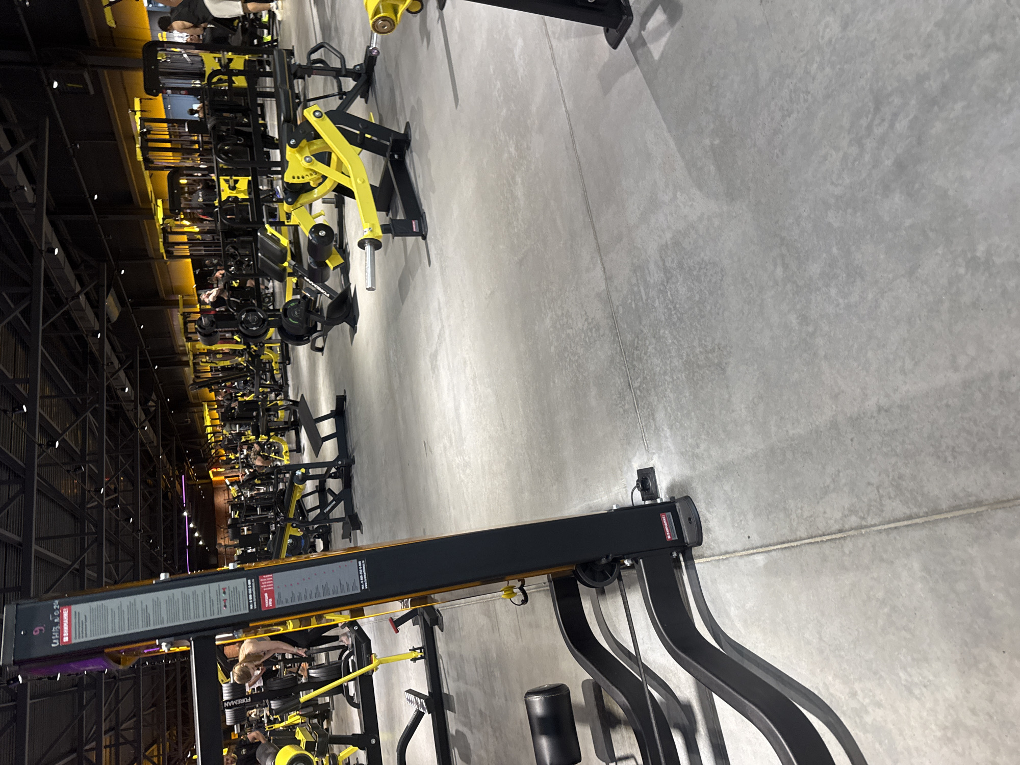 The image shows the interior of a modern gym, likely located in Komsomolsk-on-Amur, Russia. The gym features a variety of weight machines with a distinctive yellow and black color scheme. The floor is concrete. In the foreground, a large black and yellow weight machine is partially visible, with a black padded seat and a black metal frame. A tall, black vertical support with a yellow trim is on the left side, displaying instructions and advertisements. In the background, numerous other weight machines, mostly yellow and black, are arranged in rows. Several people are present in the gym, some operating the machines and others walking around. The ceiling has exposed industrial-style lighting and support structures. The overall environment suggests a functional and well-equipped fitness facility.