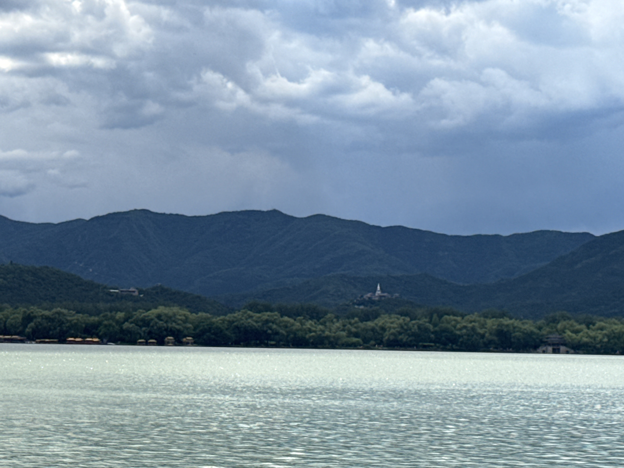 The image captures a panoramic landscape featuring a large body of water, a tree-lined shore, mountains, and a cloudy sky in Haidian District, China. The foreground is dominated by a light teal-colored body of water, exhibiting numerous small ripples across its surface and scattered points of light reflection. Directly behind the water, a dense band of dark green trees forms the immediate shoreline. Along this shoreline, several small, dark-roofed structures, possibly pavilions or boathouses, are partially visible on the left and a larger, multi-tiered building is discernible on the far right. Beyond the trees, a series of rolling, dark green mountains rise, appearing somewhat muted by distance. On a central peak in the mid-distance, a slender, white, conical pagoda or stupa with a pointed finial is prominently visible. The sky, occupying the top third of the frame, is overcast with layers of grey and white stratus clouds. No human figures or discernible actions are present in the scene.