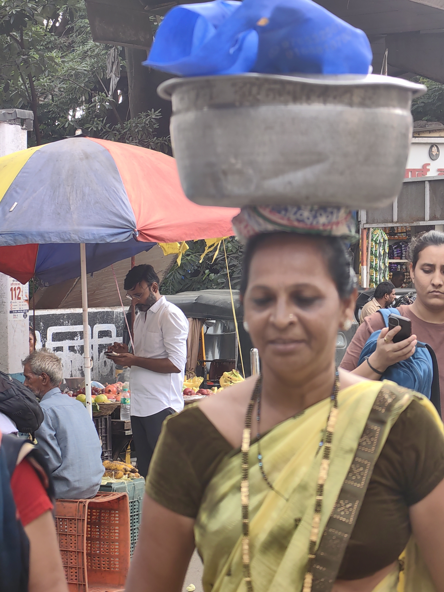 The image depicts an outdoor market scene in Mira-Bhayander, India. In the foreground, a woman is walking towards the viewer, carrying a large metal pot on her head, covered with a blue plastic bag. She is wearing a yellow sari with a dark brown blouse and a long beaded necklace. To her right, another woman, partially visible, is looking down at her mobile phone. In the middle ground, a man in a white shirt and glasses is standing under a colorful umbrella, looking at his phone. Next to him, a man with gray hair is seated, facing away from the camera. The background shows a street scene with stalls, produce, and vehicles, including an auto-rickshaw. The environment suggests a busy marketplace with food vendors.