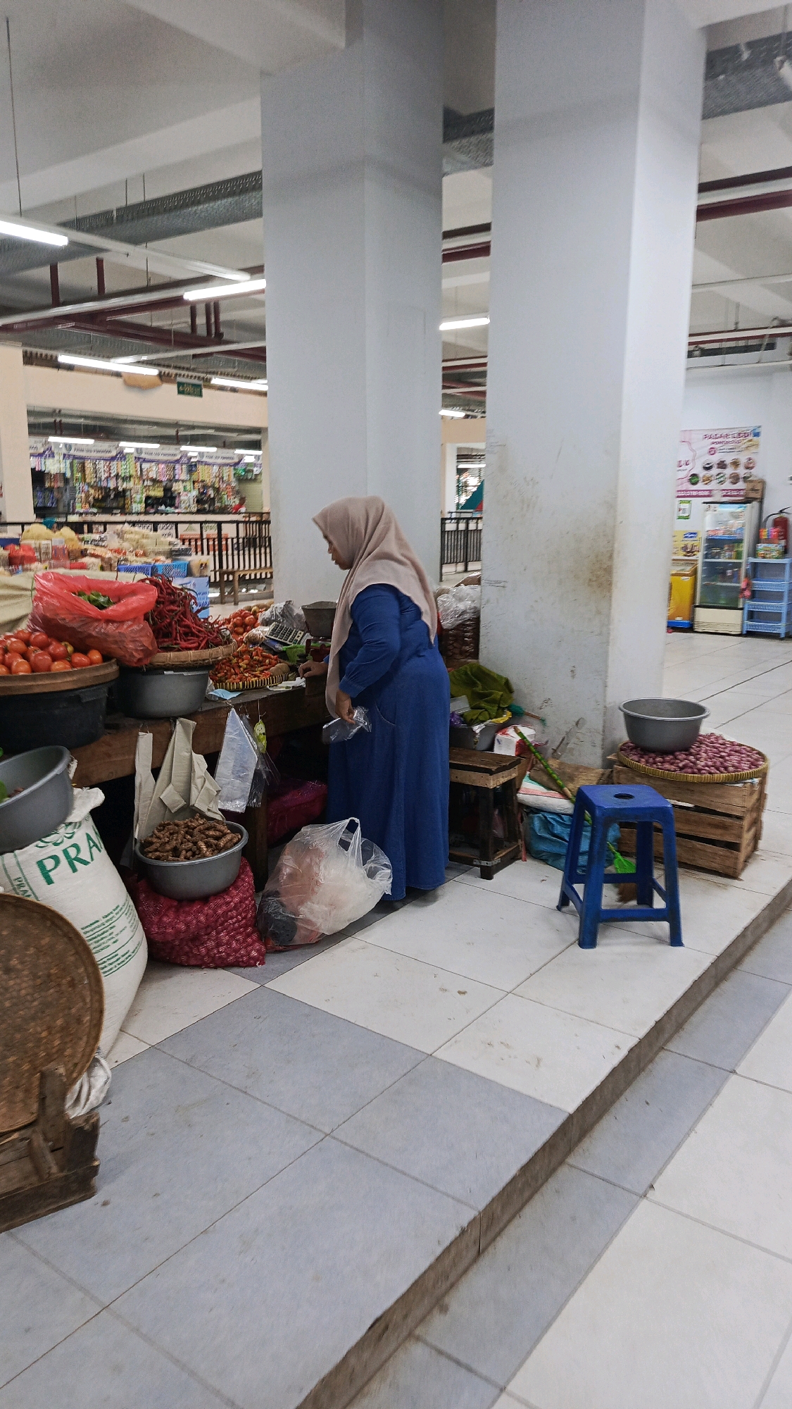 A woman wearing a hijab and a blue dress is standing at a market stall, attending to produce. She is holding a plastic bag. The stall is filled with various vegetables, including tomatoes, chilies, and onions, displayed in baskets and sacks. Behind her, shelves are stocked with packaged goods. The environment appears to be an indoor market or a supermarket, with tiled flooring and bright overhead lighting. A blue plastic stool is positioned near the stall. The scene suggests a commercial setting within the city of Ponorogo, Indonesia.