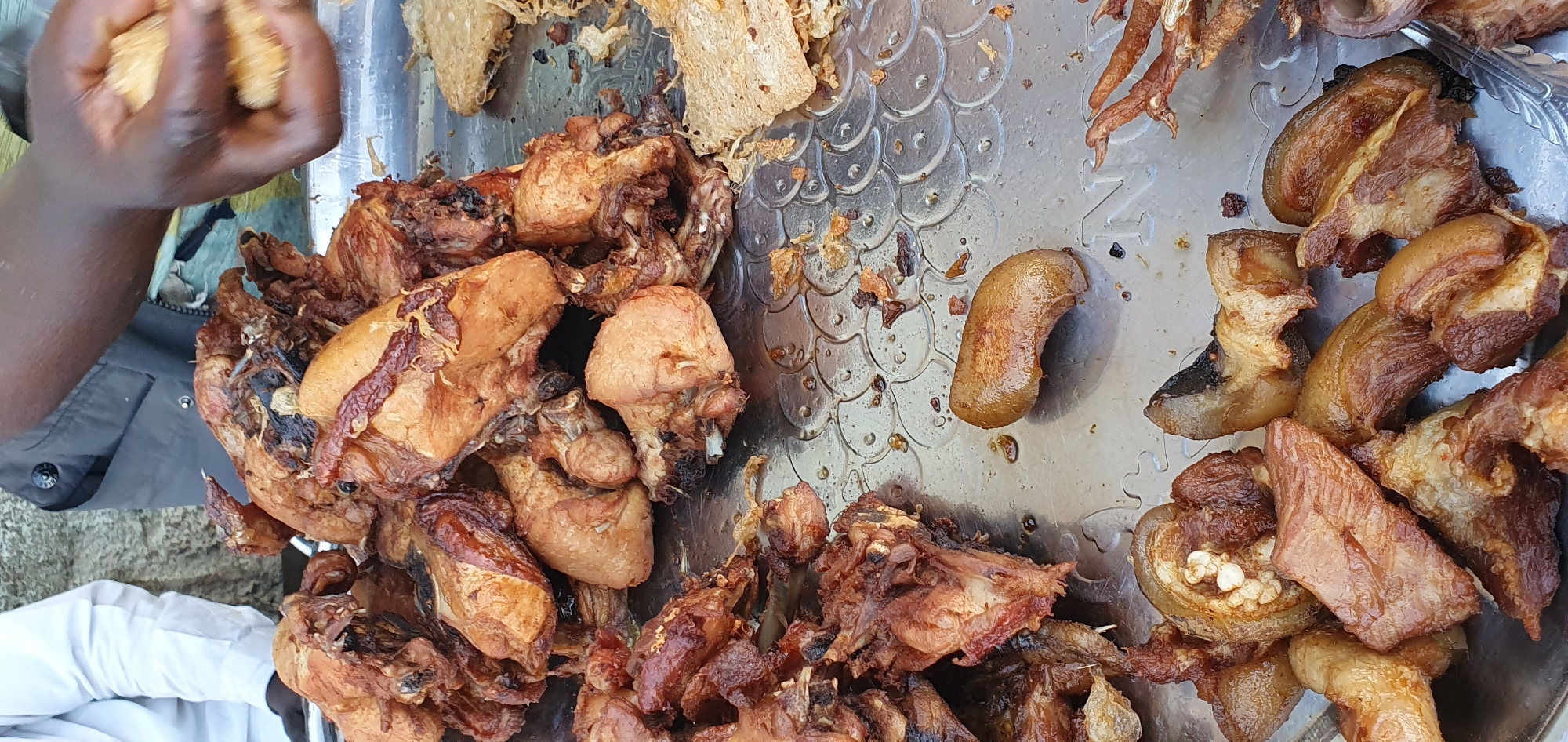 A hand is visible on the left side of the image, holding pieces of fried chicken. The majority of the image is filled with a large platter of fried chicken pieces and some other fried meats, possibly pork. The chicken pieces are of various sizes and appear to be seasoned and fried to a golden brown. Some pieces show visible bones. The platter is made of a shiny, metallic material with an embossed pattern that resembles scales. Scattered around the food are small pieces of what appears to be dried food or seasoning. In the upper left corner, a piece of what looks like fried plantain or yam can be seen. The background is out of focus but suggests an outdoor setting, with hints of clothing and possibly a table. The overall impression is of a street food vendor or a casual meal preparation. The lighting suggests daytime. The location context is Bukuru, Nigeria. No visible text is present in the image.