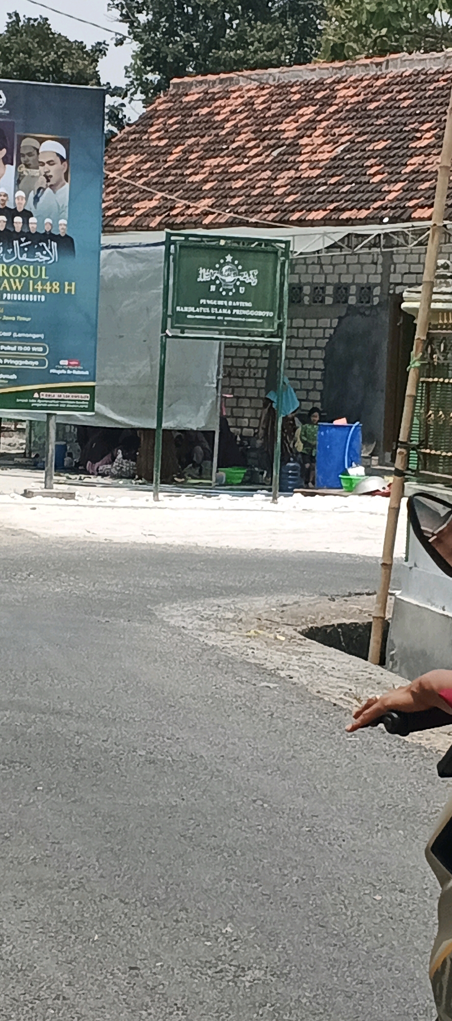The image shows a street scene in Kanugrahan, Indonesia. In the foreground, a person is seen from the chest down, with their hand resting on a black handlebar, suggesting they are on a motorcycle or similar vehicle. Their left hand is visible, with pink nail polish.
In the middle ground, there is a street with a curb. Behind the curb, there is a temporary structure covered with a white tarp, set up under a green sign. The sign, partially obscured, displays Arabic script and an emblem, possibly indicating a religious or community organization. Underneath the tarp, several people are seated or lying down, some appearing to be sleeping or resting. Various objects, including green buckets, plastic containers, and a large blue barrel, are present around the structure. The ground beneath the tarp is covered with mats and blankets.
Further in the background, there are buildings with tiled roofs and brick walls. A large banner is visible on the left side of the image, featuring images of people in religious attire and text in Arabic and Indonesian. The banner mentions "ROSUL AW 1448 H" and "PRINGGOTOYO". Trees with green foliage are seen above the buildings. The overall environment appears to be a daytime scene in a moderately urban or semi-rural area.