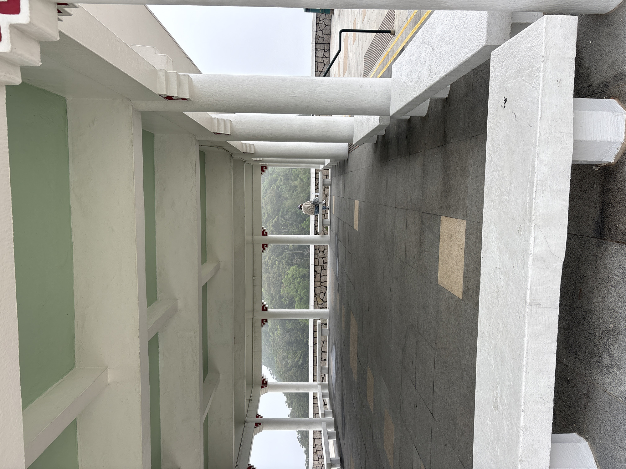 The image depicts a covered walkway in what appears to be a park or public space in Hong Kong, China. The walkway is constructed with white pillars and beams, creating a series of framed views of the surrounding landscape. A person sits on a bench further down the walkway. The ground is paved with dark gray tiles with occasional light brown tiles. The structure is painted white with light green accents. The view from the walkway reveals a green, hilly landscape partially obscured by mist.