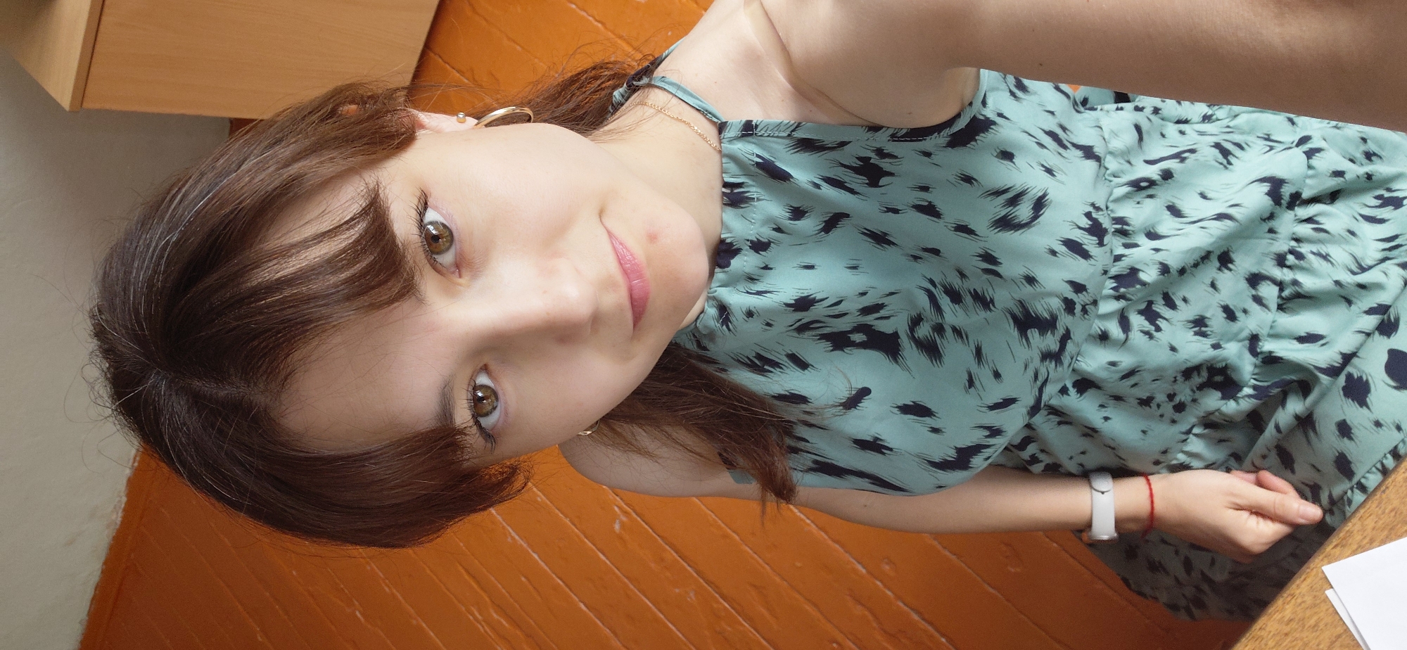 The image shows a young woman lying down on an orange wooden floor, looking up at the camera. She has long, brown hair with some lighter streaks, and is wearing a light teal dress with a black abstract pattern. She is also wearing a thin gold necklace and a gold hoop earring in her visible ear. On her left wrist, she has a white fitness tracker and a thin red string bracelet. Her expression is a slight, gentle smile. The background shows a light-colored wooden cabinet or desk and the textured orange floor. The lighting suggests it is daytime, possibly indoors. No text is visible in the image. The overall mood is casual and relaxed.