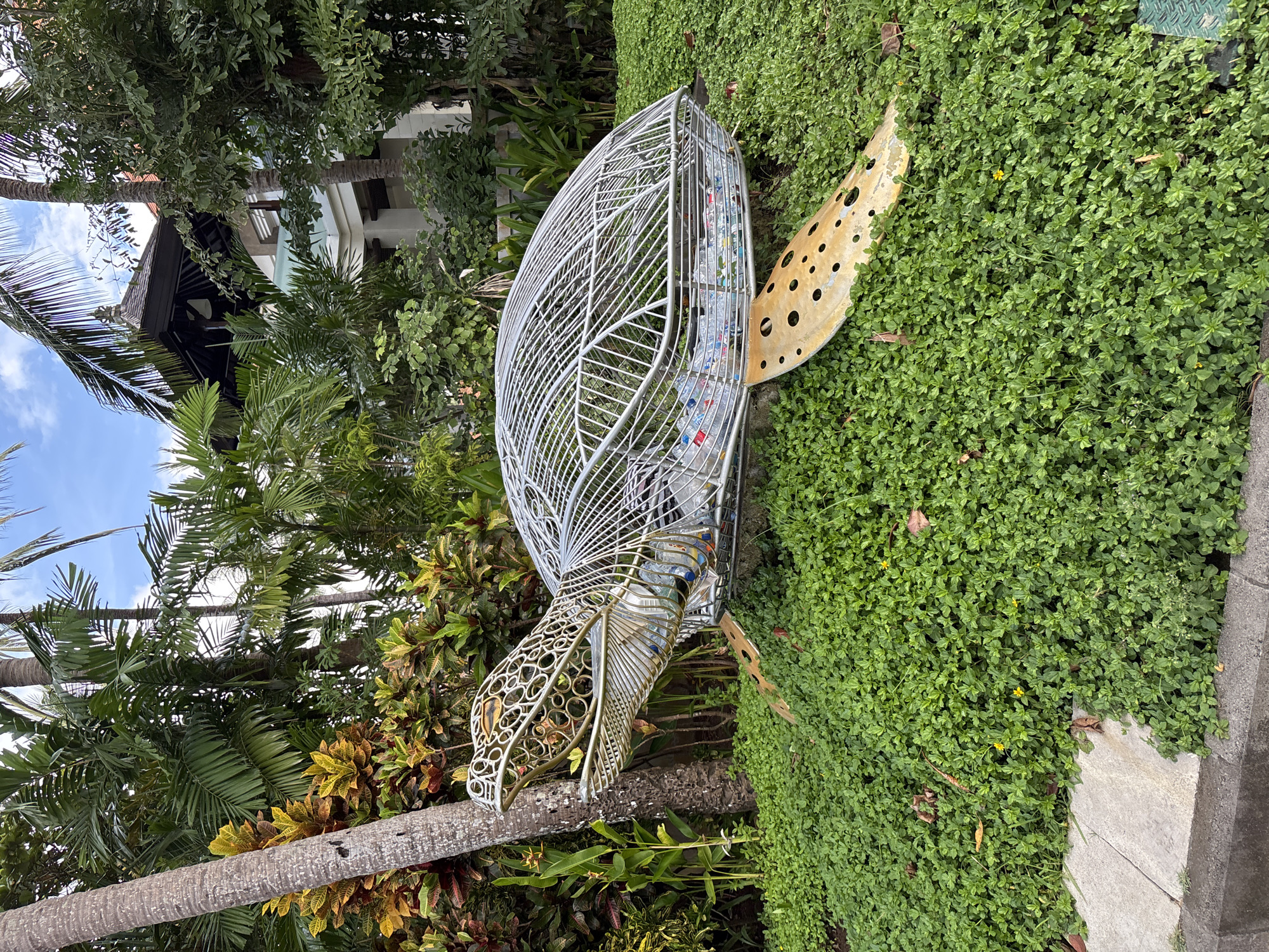 The image shows a wire-frame sculpture of a sea turtle in a grassy area in Kuta Selatan, Indonesia. The turtle's shell is made of white wire and is filled with clear plastic bottles. Its head, legs, and tail are made of yellow metal, with circular cutouts visible on the tail. Lush tropical vegetation, including palm trees, surrounds the sculpture. A portion of a building can be seen in the background, partially obscured by the trees. A concrete sidewalk or curb is visible on the right side of the frame, bordering the grassy area where the sculpture is placed.