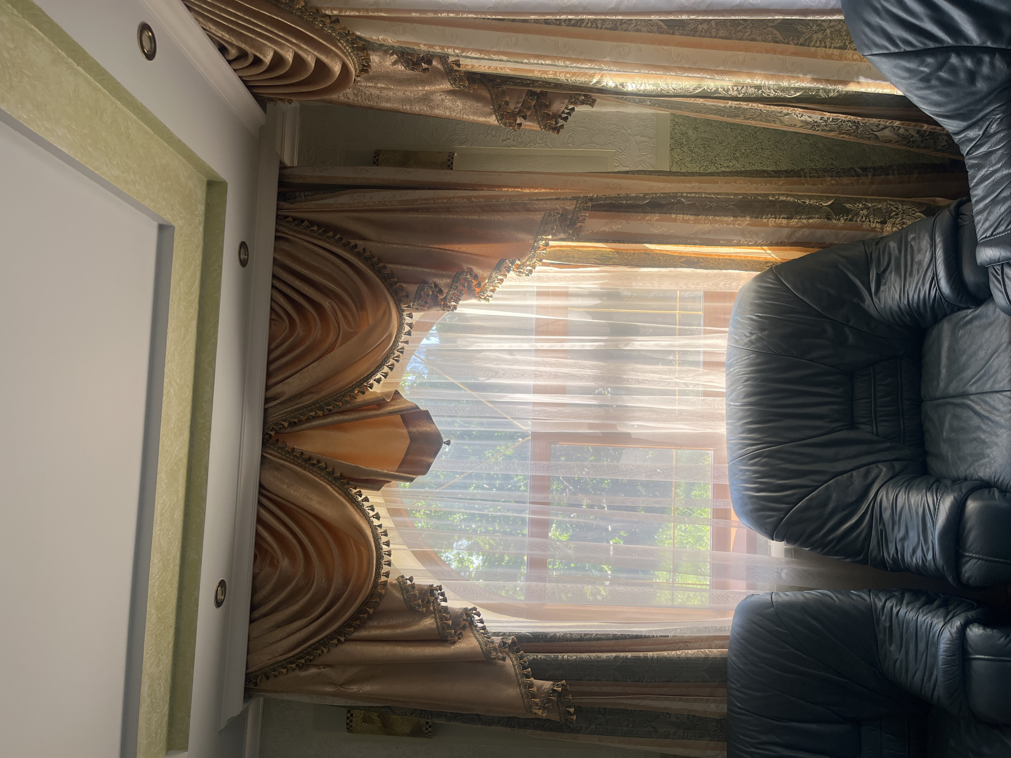 In the city of Tarnivtsi, Ukraine, this image captures an indoor scene featuring a large arched window with ornate golden drapes. The drapes are layered, with a sheer inner curtain allowing natural light to filter through, illuminating lush green foliage visible outside. Two dark leather armchairs are positioned in the foreground, facing the window. The ceiling features recessed lighting and decorative molding, one section of which is painted a light green. The walls appear to be a muted green, with subtle textured wallpaper. The overall impression is one of a well-decorated room with a focus on natural light and comfortable seating.