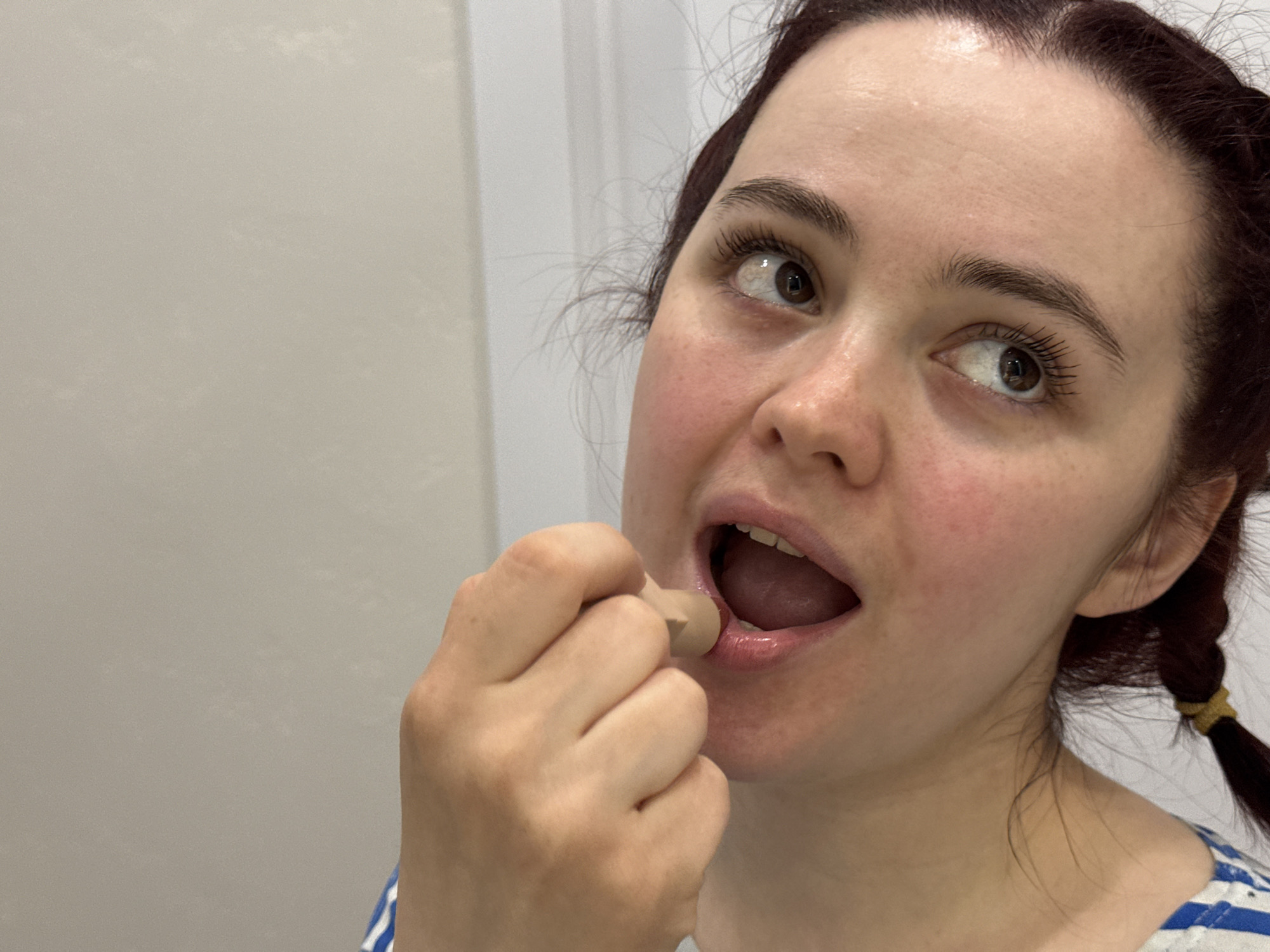 A close-up, candid shot depicts a young woman engaged in personal grooming indoors, in Izhevsk, Russia. The woman, shown from the chest up, has fair skin with some natural redness on her cheeks and forehead. Her dark hair, possibly reddish-brown, is pulled back, with a braid secured by a yellow hair tie visible on the right side of the frame. Her eyebrows are dark and well-defined, and her eyelashes appear long, possibly enhanced with mascara.
She is seen holding a small, light brown, soft applicator, likely a makeup sponge or balm applicator, between her thumb and forefinger, positioned near her open mouth. Her mouth is open wide, revealing her tongue and the inside of her mouth, suggesting she is in the process of applying a product to her lips or teeth. Her dark brown eyes are wide open and directed upwards and to the right, conveying an expression of focus, surprise, or perhaps looking at her reflection in a mirror. She wears a top with blue and white stripes.
The background is a plain, light-colored wall, possibly beige or off-white, with a hint of a white doorframe visible on the far right, indicating an indoor setting like a bathroom or bedroom. The lighting is bright and even, consistent with an indoor environment during daytime, though no specific time of day can be determined. There is no visible text in the image.