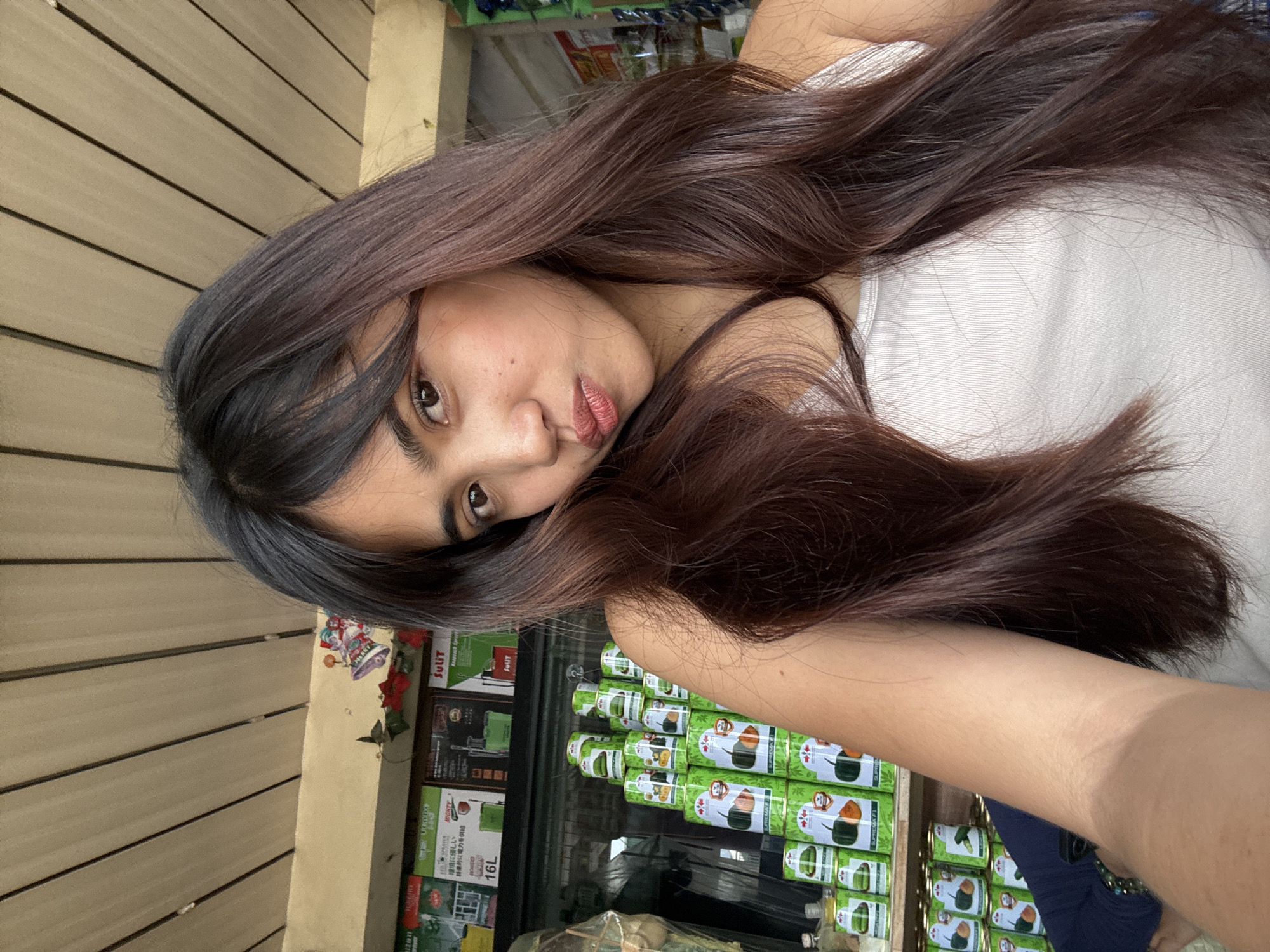 A young woman with long, dark brown hair, styled with bangs, takes a selfie in what appears to be a small store or market stall. She is wearing a white ribbed tank top and has her lips pursed. In the background, shelves are stocked with various products, including green cans with images of vegetables and promotional posters. Above her, a wooden slatted ceiling is visible, with festive Christmas decorations hanging. The environment suggests a local business setting, possibly in Tagum City, Philippines, given the context.