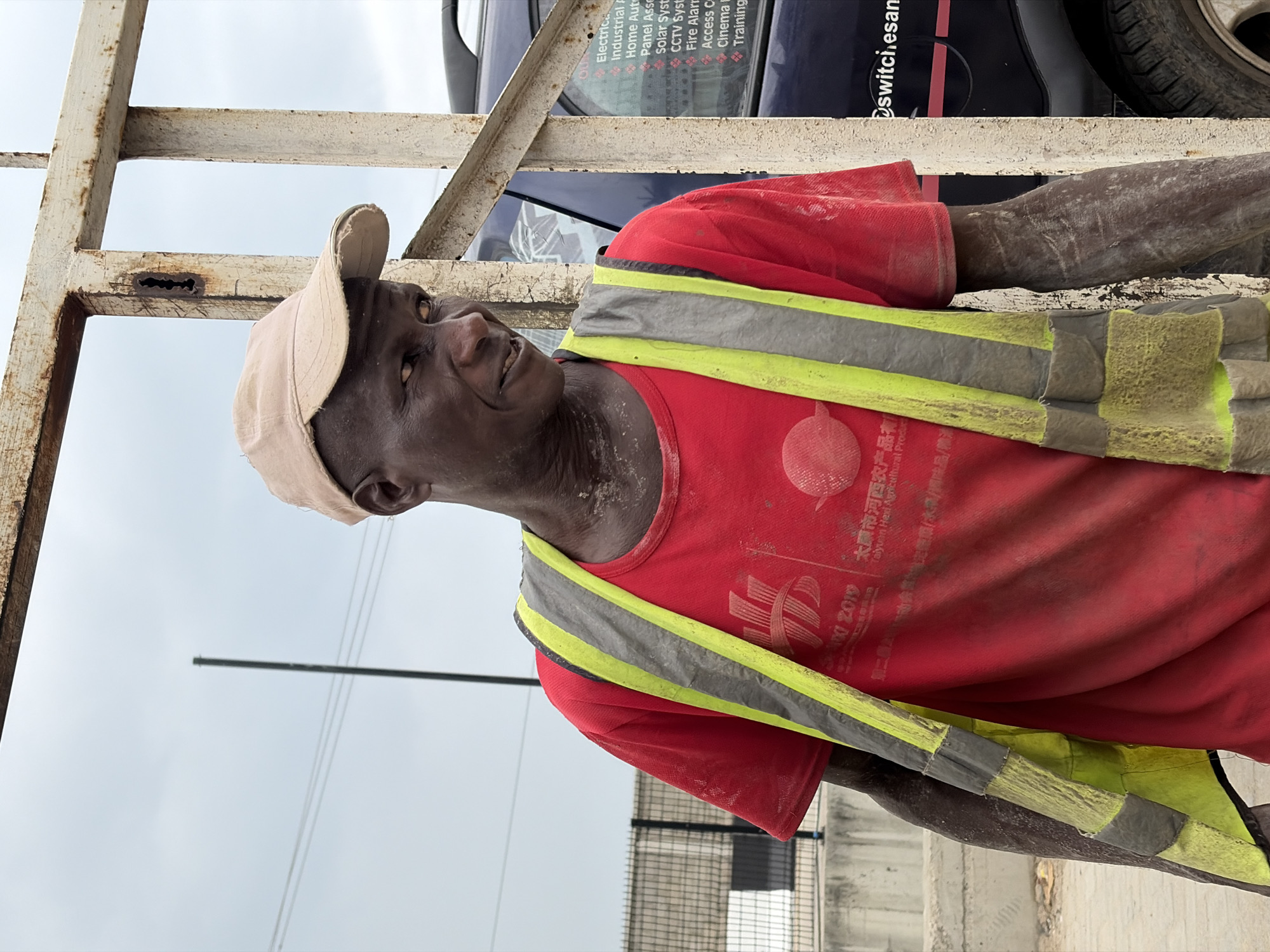 A construction worker wearing a red t-shirt, reflective vest, and tan cap is seen in Igboefon, Nigeria. He is standing near a metal structure, with a vehicle visible behind him. The vehicle has text on the side indicating it's related to electrical, industrial, and home automation services, including CCTV systems and fire alarms. The t-shirt has Chinese characters and the text "Taiyuan Hexi Agricultural Product Exhibition 2019." The overall scene appears to be an outdoor environment.