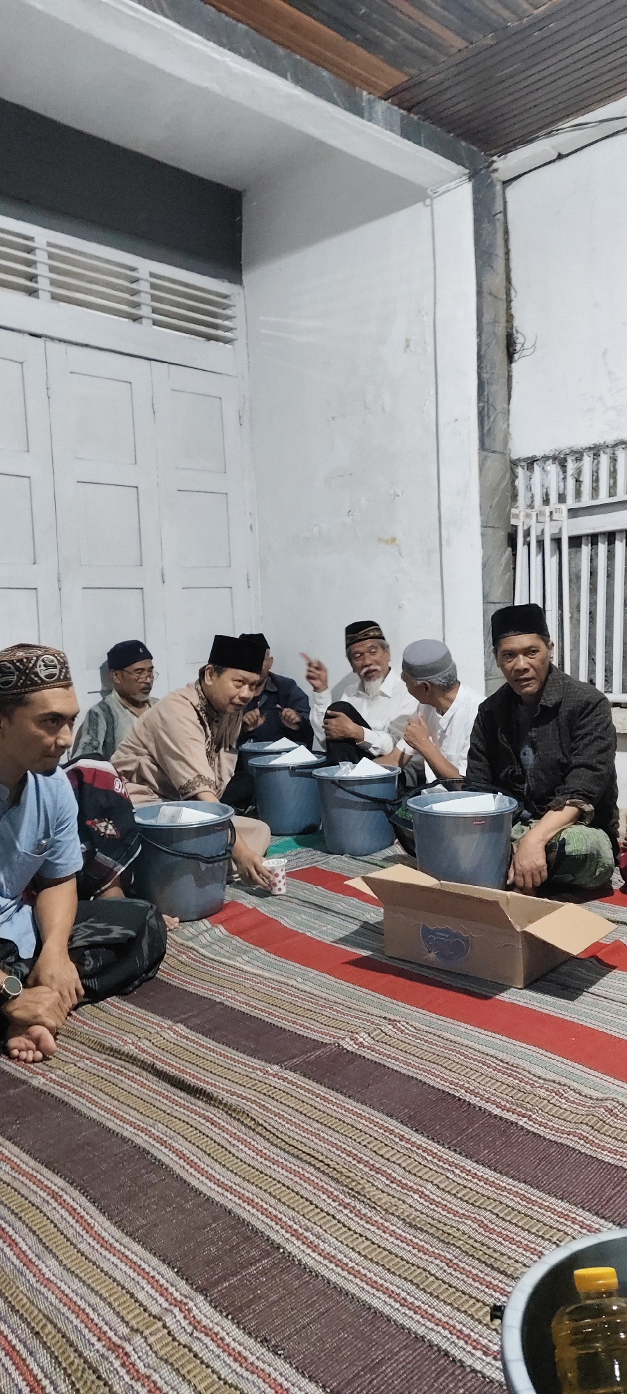 The image depicts a gathering of at least seven men, all wearing traditional Islamic caps, sitting on a woven mat on the ground. The setting appears to be an outdoor or semi-outdoor area, possibly a courtyard or veranda, adjacent to a white wall with a large, white louvered door or window. The lighting suggests it is evening or night.
Several men are engaged in conversation or listening, with one man in the center actively gesturing. In front of the men are several large, blue plastic buckets, some containing white paper or objects. A cardboard box, partially open, sits on the mat to the right. A bottle of yellow liquid, possibly oil, is visible in a dark container in the foreground to the far right. The mat is a rectangular woven surface with horizontal stripes of varying colors, including dark brown, beige, red, and lighter brown. The overall atmosphere suggests a casual, communal gathering.
Given the context and typical attire, this scene could plausibly be located in an area with a strong Muslim population in Malang, Indonesia, such as a residential neighborhood or a religious center. However, without specific landmarks or identifying features, a precise location within the city of Malang cannot be determined from the image alone.