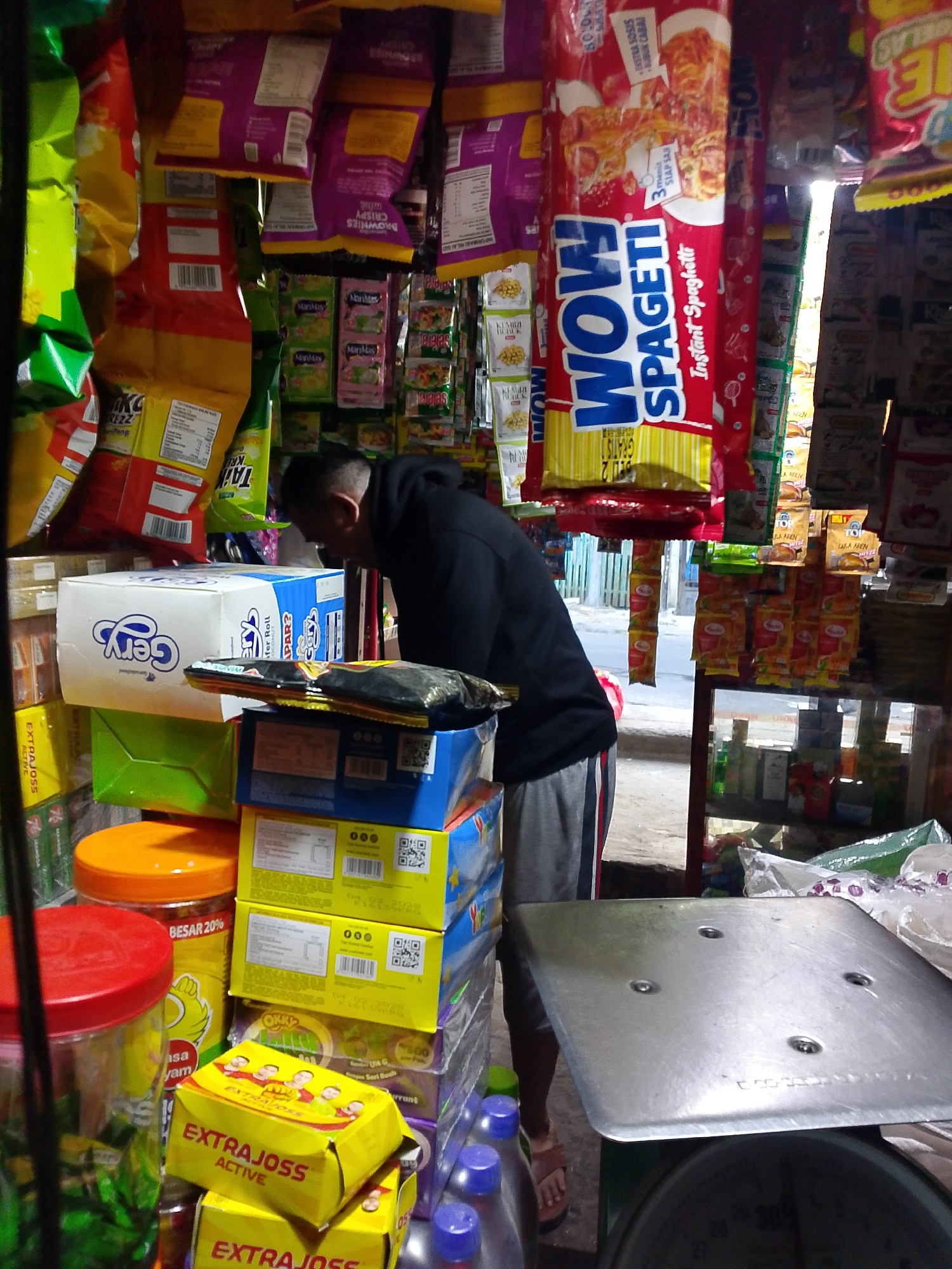 This is an eye-level, medium shot of a small, open-air convenience store, possibly a warung, located on a street in Palembang, Indonesia. The store is densely packed with various food and drink items. In the foreground, a man, wearing a black hooded sweatshirt and grey sweatpants, is bending down, looking at boxes stacked on a shelf. He appears to be a vendor or a customer. To his left, there are large clear plastic containers with red and orange lids filled with colorful snacks and other items. In the center, a stack of yellow and blue boxes are visible, some with labels indicating they contain "Gery" products and "Extra Joss" energy drinks. Hanging from the ceiling are numerous brightly colored snack bags, including red bags of "Wow Spaghetti" and purple bags of "Brownies Crispy." The store is illuminated by natural light coming from the background, which appears to be a street scene with some buildings and a bright sky. The overall impression is of a vibrant and busy local market setting.