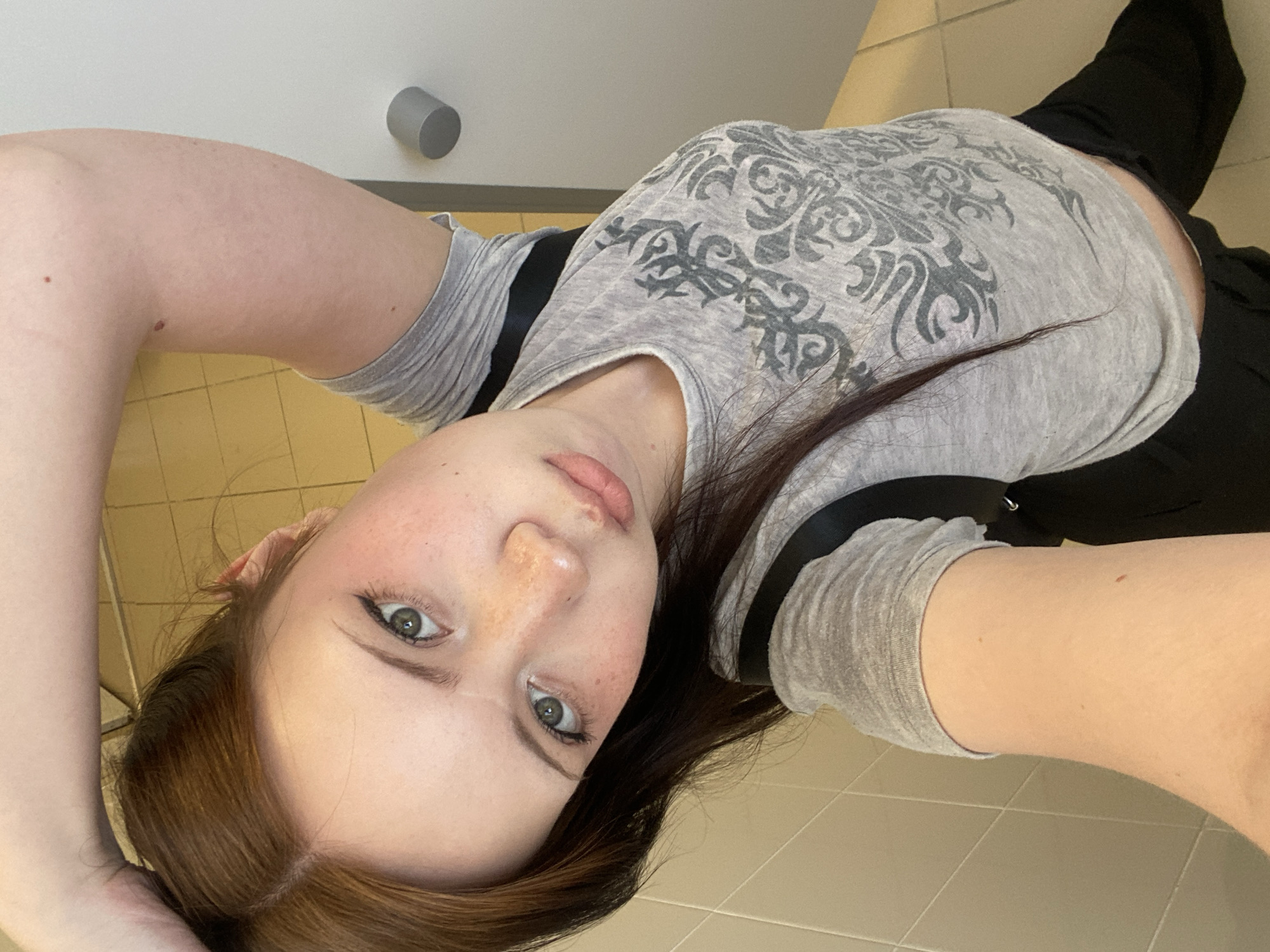 The image depicts a young woman in a gray t-shirt with a dark pattern, and black pants, lying down on a light-colored tile floor. Her head is tilted, and she is looking at the camera. A white wall with a cylindrical object attached is visible in the background. The location is Unknown, Unknown.