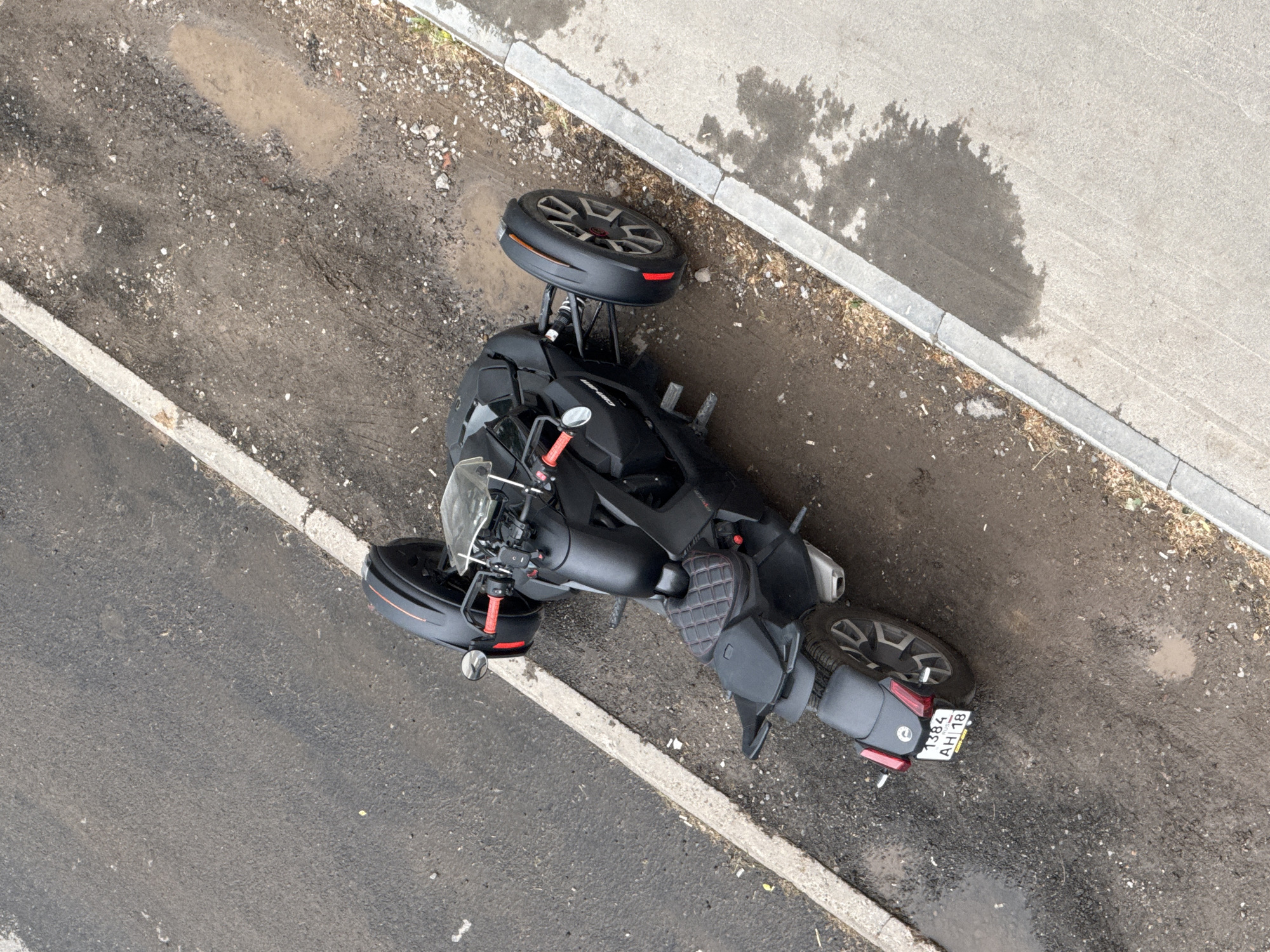 An aerial shot shows a black Can-Am Ryker three-wheeled motorcycle lying on its side in Votkinsk, Russia. The vehicle is near a curb with visible damage, suggesting it may have fallen or been intentionally tipped over. The license plate is partially visible and reads "1384 AH 18". The area around the vehicle shows signs of wear and tear, with cracked asphalt, dirt, and small puddles. There is no visible human presence.