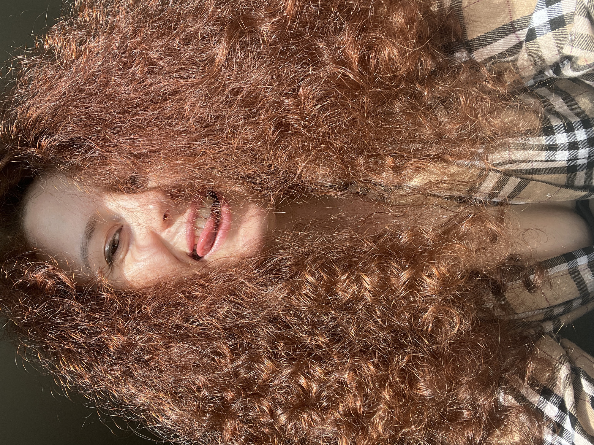 A close-up shot, likely taken with a cell phone, shows a woman with reddish-brown, curly hair. Her face is partially obscured by her hair, but one eye, part of her nose, her mouth, and tongue are visible. She appears to be lying down. A plaid shirt or blanket is visible in the background. There is no visual information in the image that would identify this as Saint Petersburg, Russia.